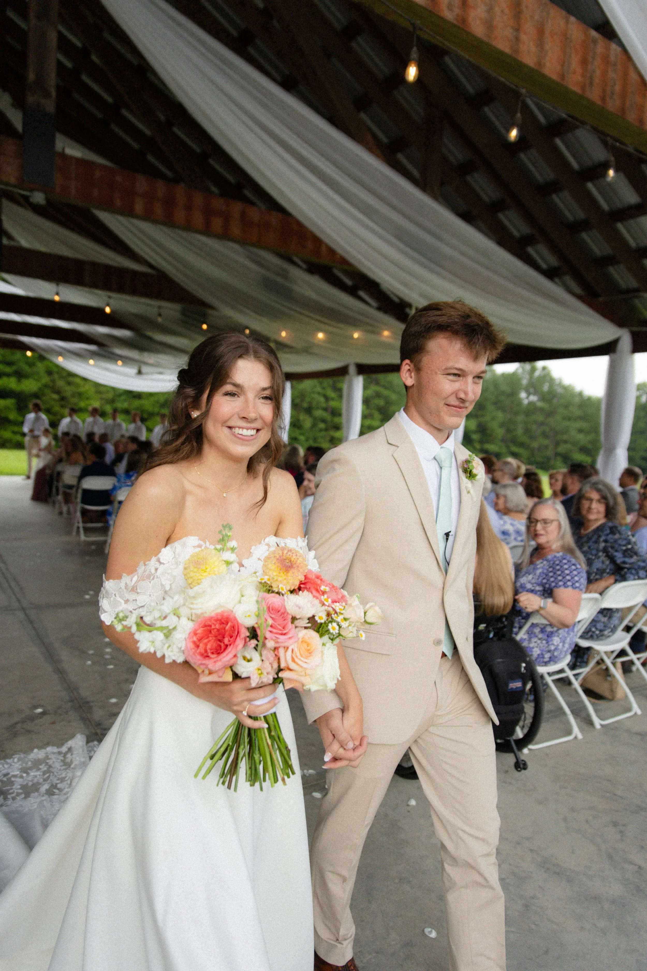 A bride and groom walking hand in hand at their outdoor wedding reception, with guests seated at tables under a covered pavilion with draped white fabric and string lights.