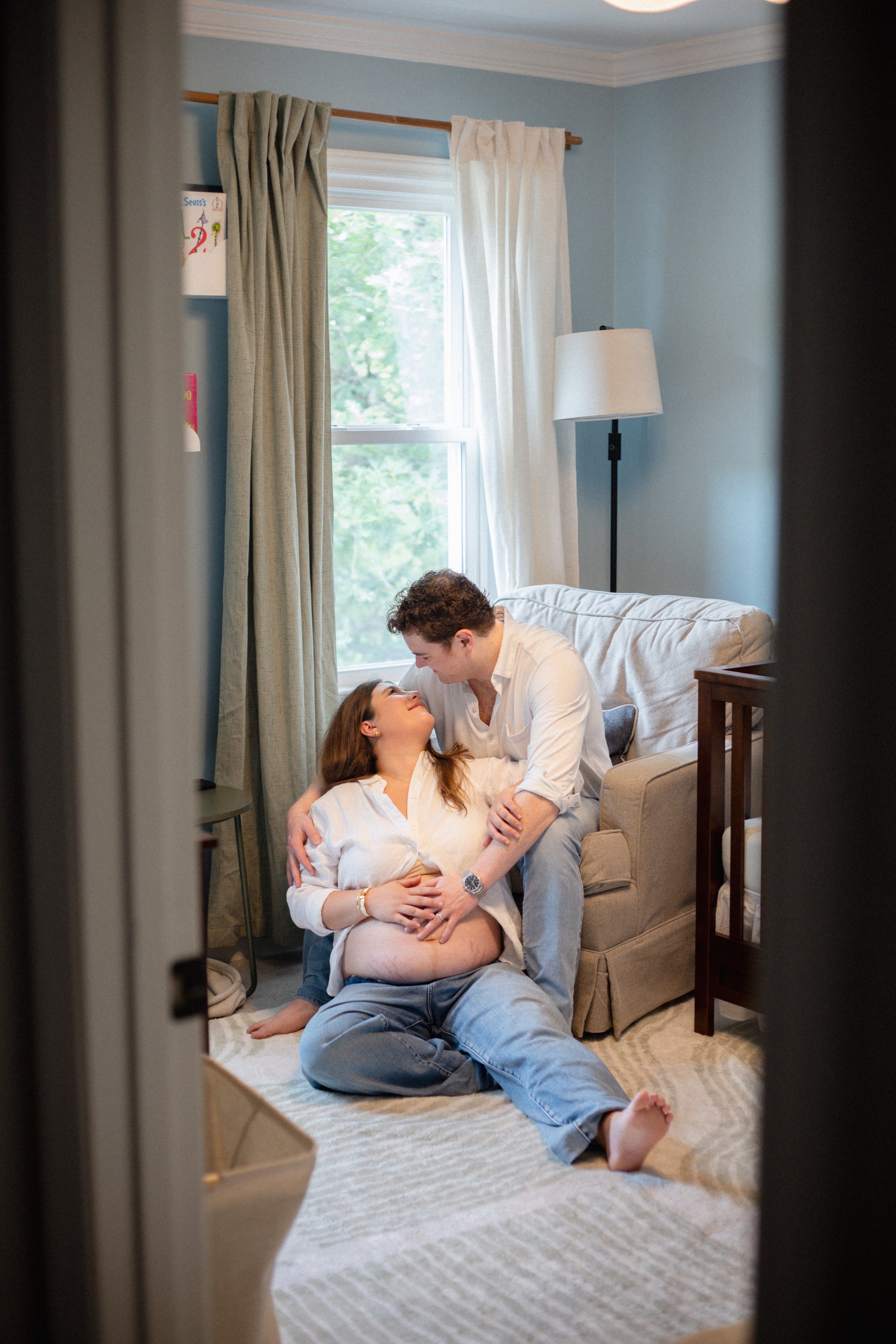 A couple is in a living room, with the man sitting on the floor and the woman lying on a couch. They are gazing into each other's eyes, and the man is leaning over the woman, who has her arms around him. The room has a window with curtains, a lamp, and some wall decorations.
