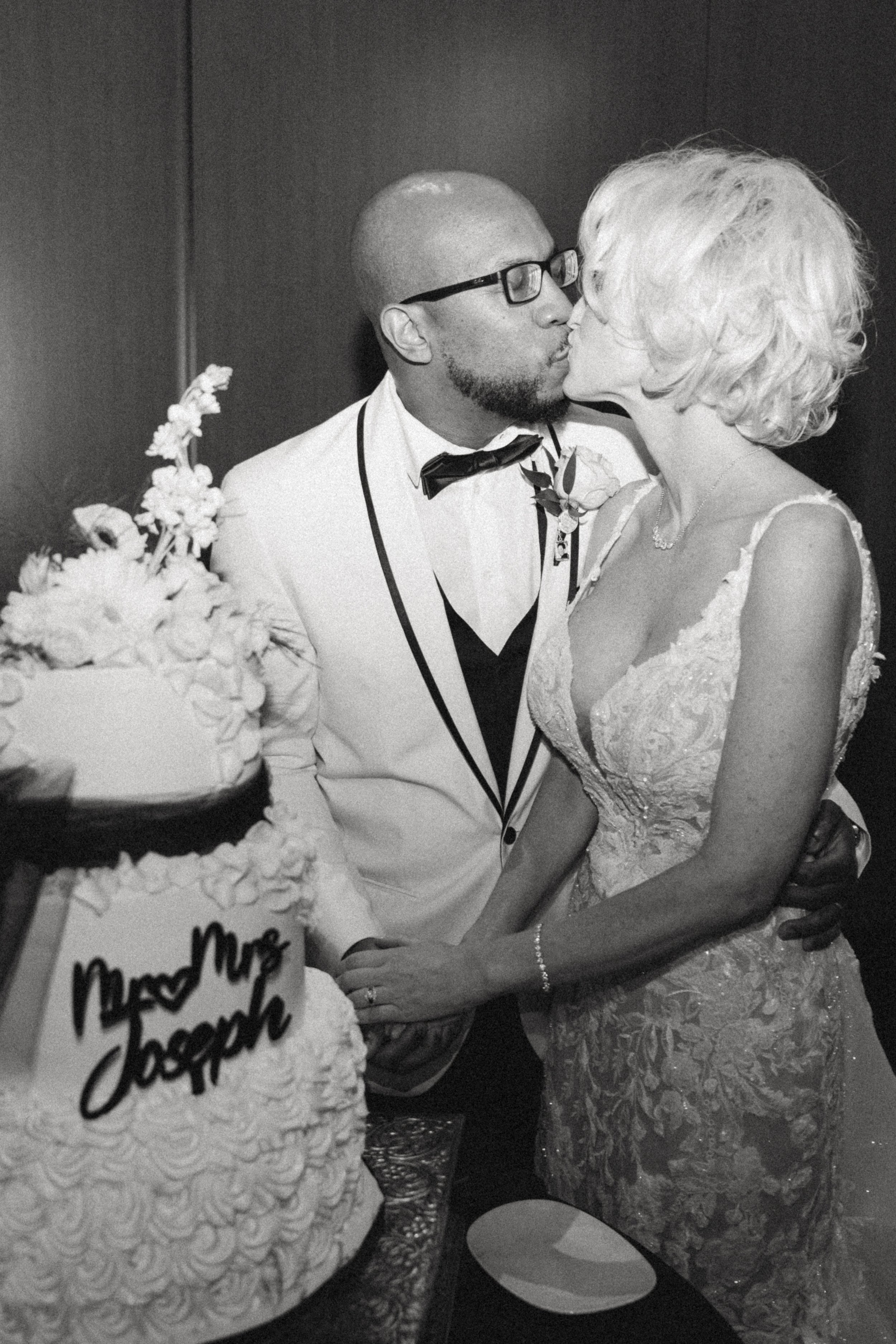 A man with glasses and a beard wearing a tuxedo is kissing an older woman in a wedding dress, with a wedding cake and flowers nearby.