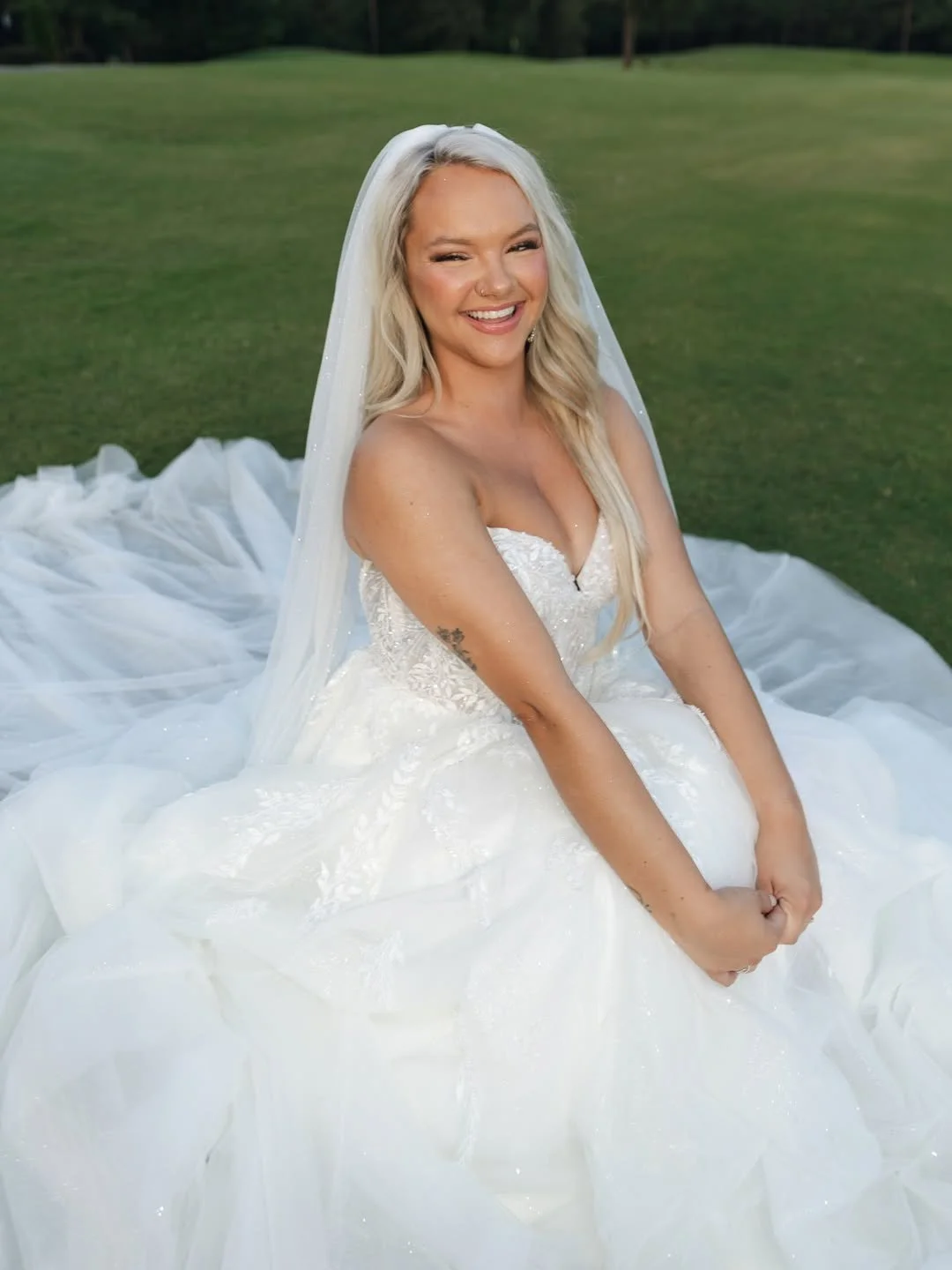 A smiling woman in a white wedding dress and veil sitting on a grassy field.