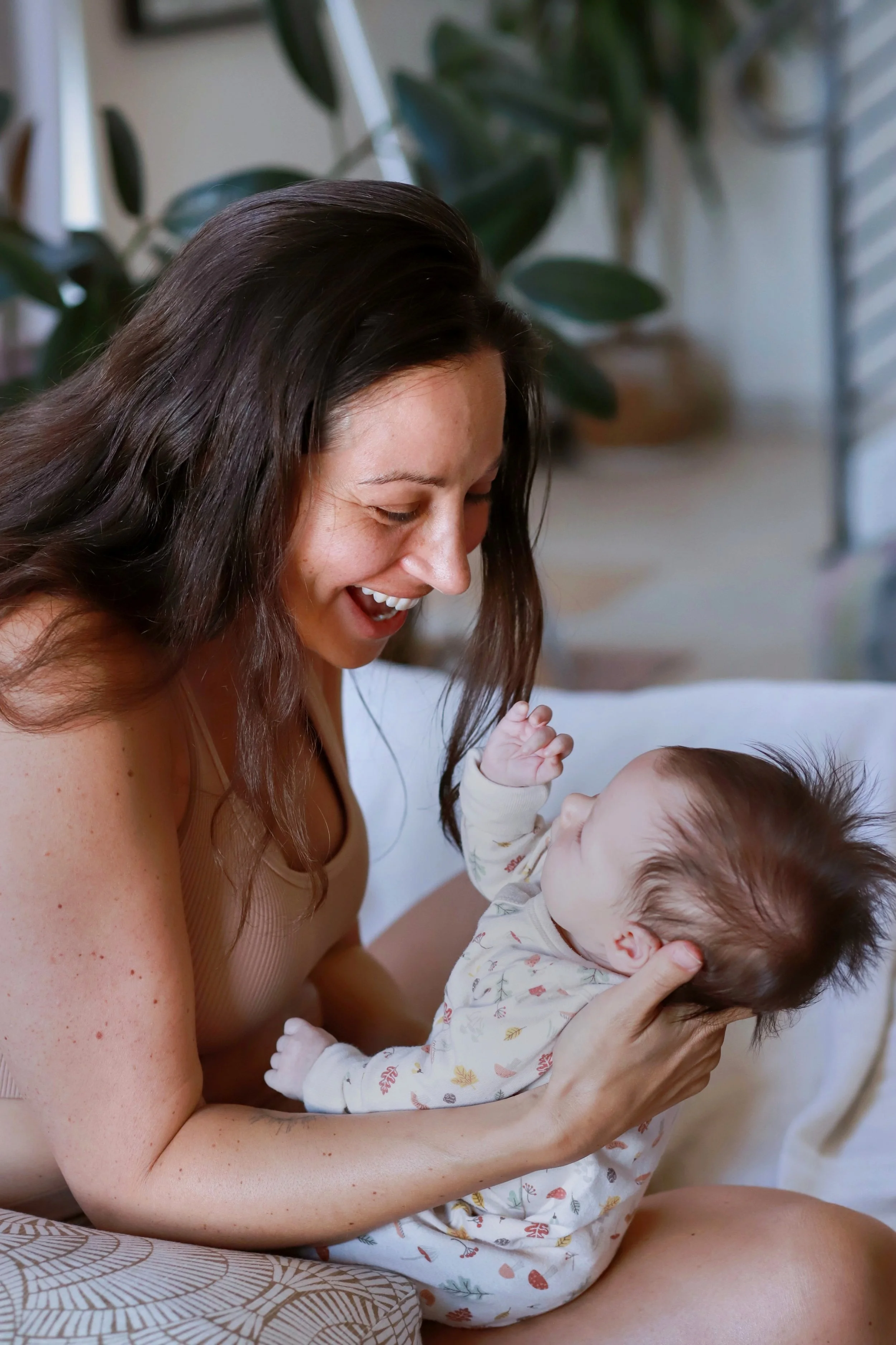 A woman with long dark hair smiling and holding a baby with spiky hair, on a white bed with a large leafy plant in the background.