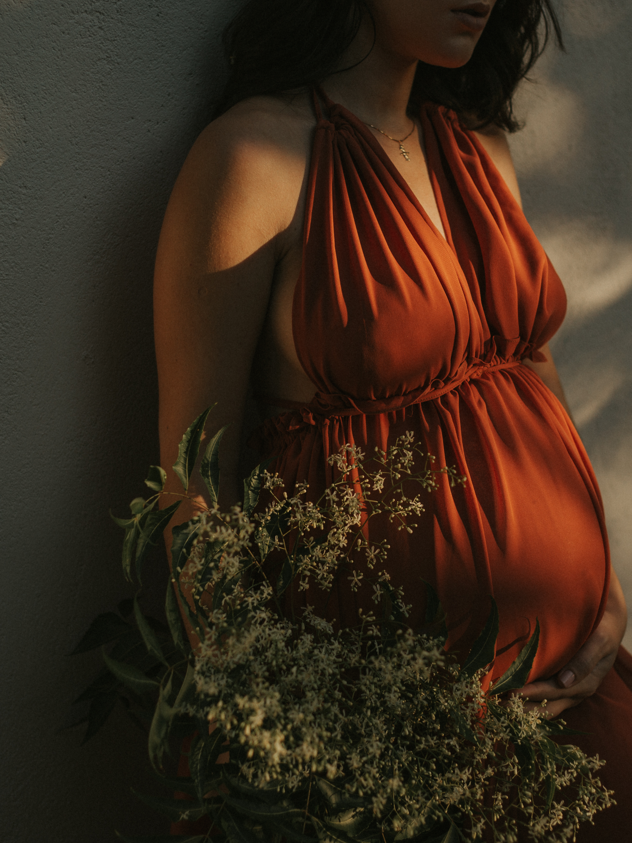 A woman in an orange dress with a floral arrangement, standing against a textured wall.
