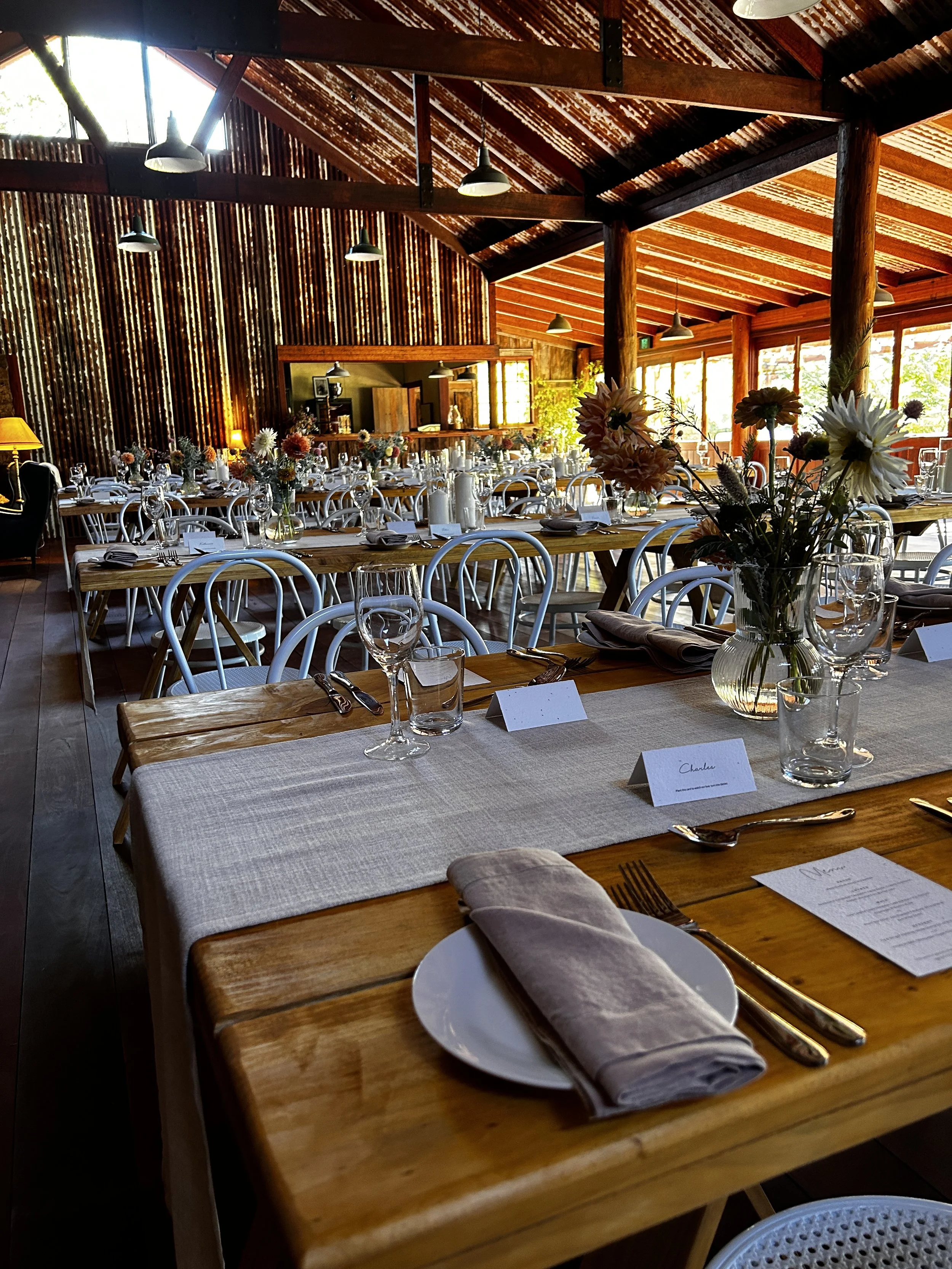 Table setting with placemats and flowers on bespoke wooden tables and white chairs in a country barn