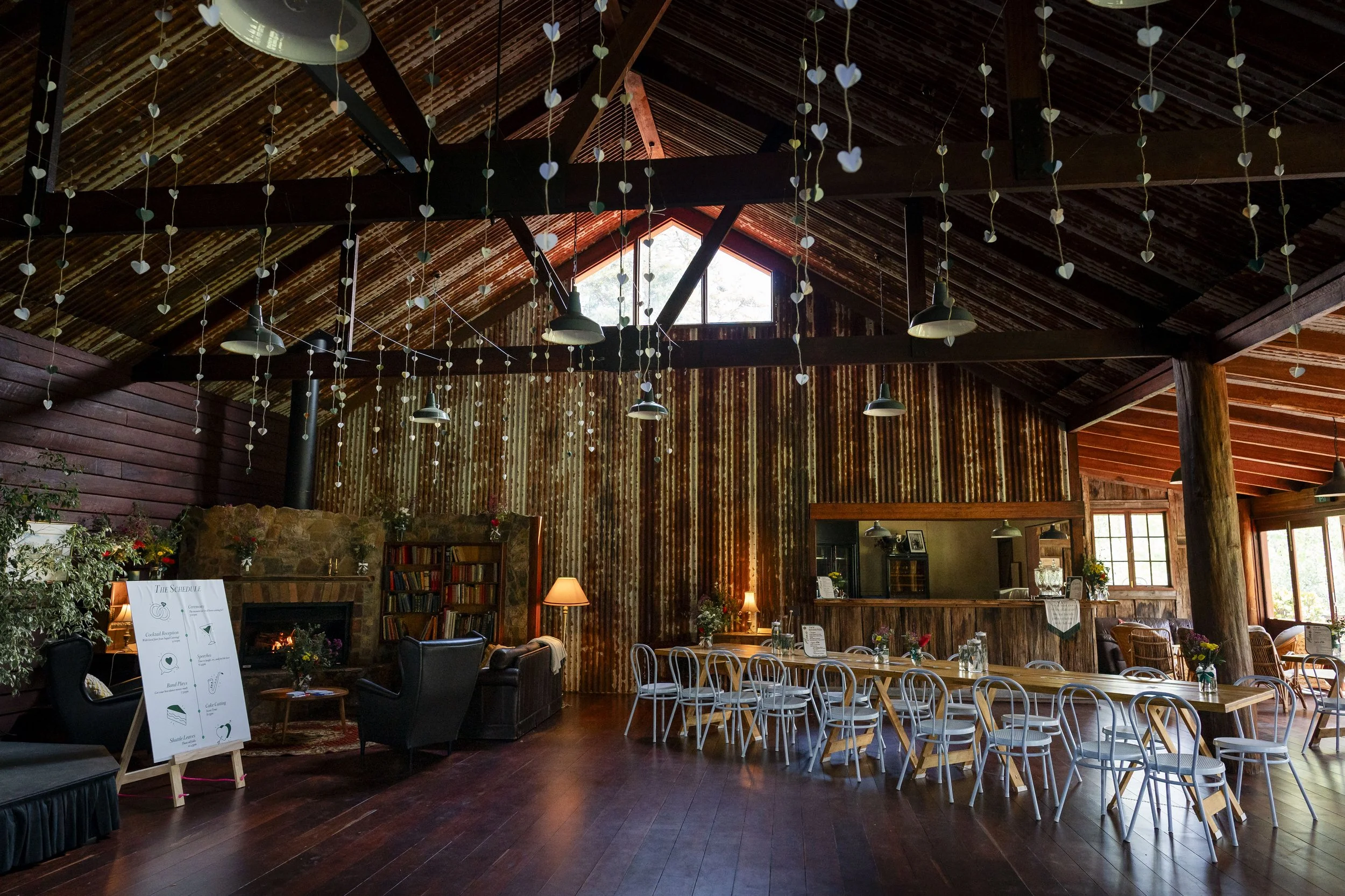 Country style barn setting with polished wooden floors and rusty corrugated walls set up tables and chairs for a wedding.  Shows a stone fire place and a dance floor
