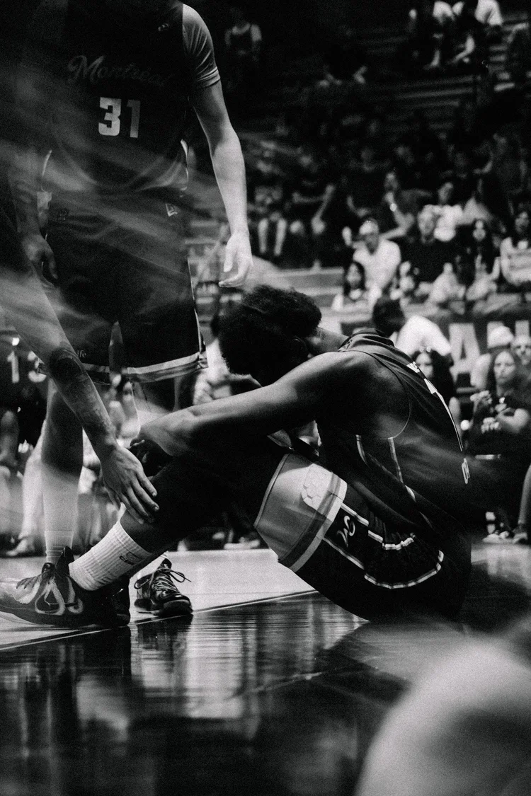 Photographe sportif Montréal - anais fabra.
Deux joueurs de basketball en action, un est assis par terre avec un genou plié, l'autre est debout, tous deux en tenue de sport, dans une salle bondée.