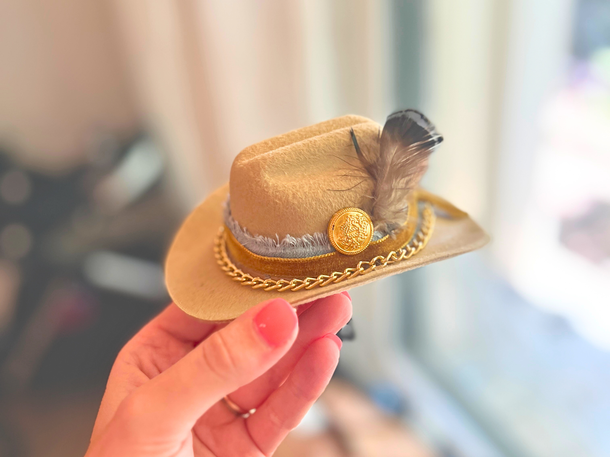 Close-up of a mini hat bar style pet cowboy hat in beige felt. Features a detailed blue ribbon trim, tiny decorative feather, and a shiny gold chain link necklace detail for small pets.