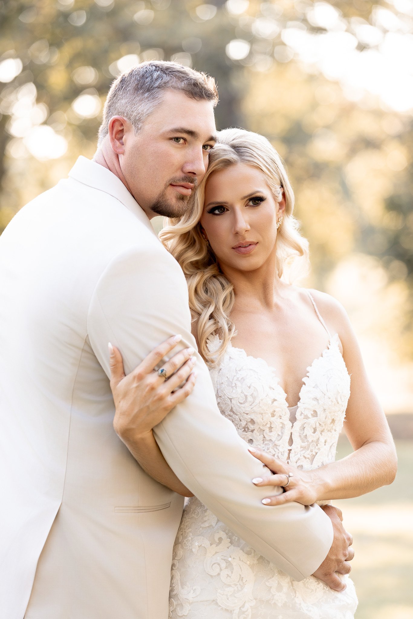 A couple, dressed in wedding attire, stands close outdoors during golden hour. The man wears a white suit and the woman wears a lace wedding dress with blonde hair styled in loose waves.
