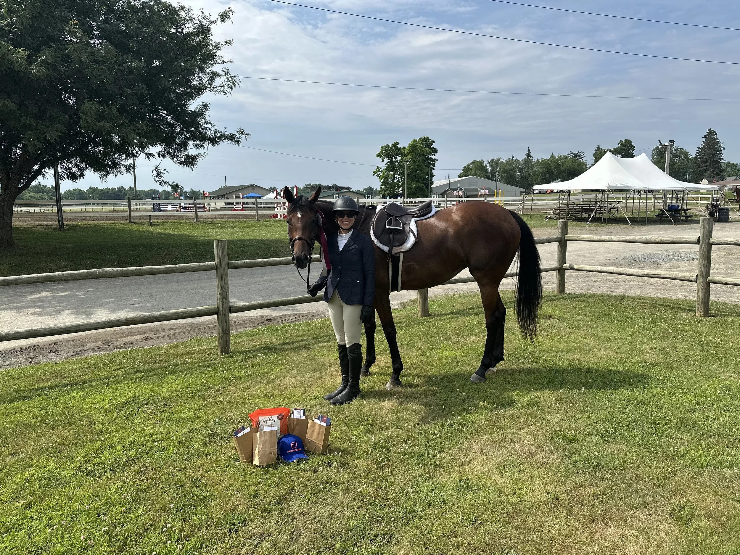 A woman in equestrian attire standing next to a saddled brown horse on a grassy area near a fence at a horse riding arena. There are prizes and items on the ground near them, and tents and structures in the background on a partly cloudy day.