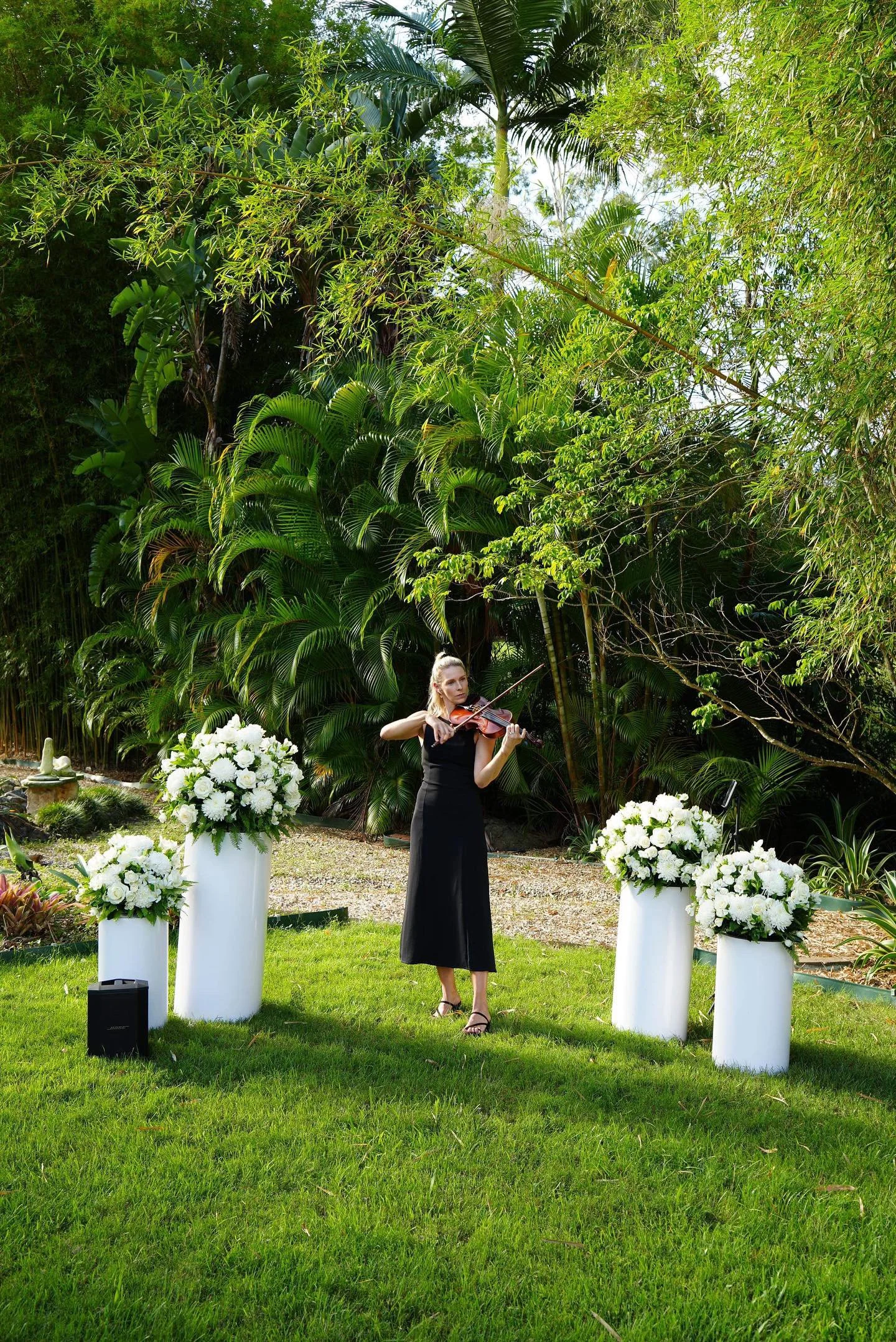 Lauren Bradnam Violinist in black dress playing violin outdoors, surrounded by white flower arrangements on white pedestals, lush green trees in the background.