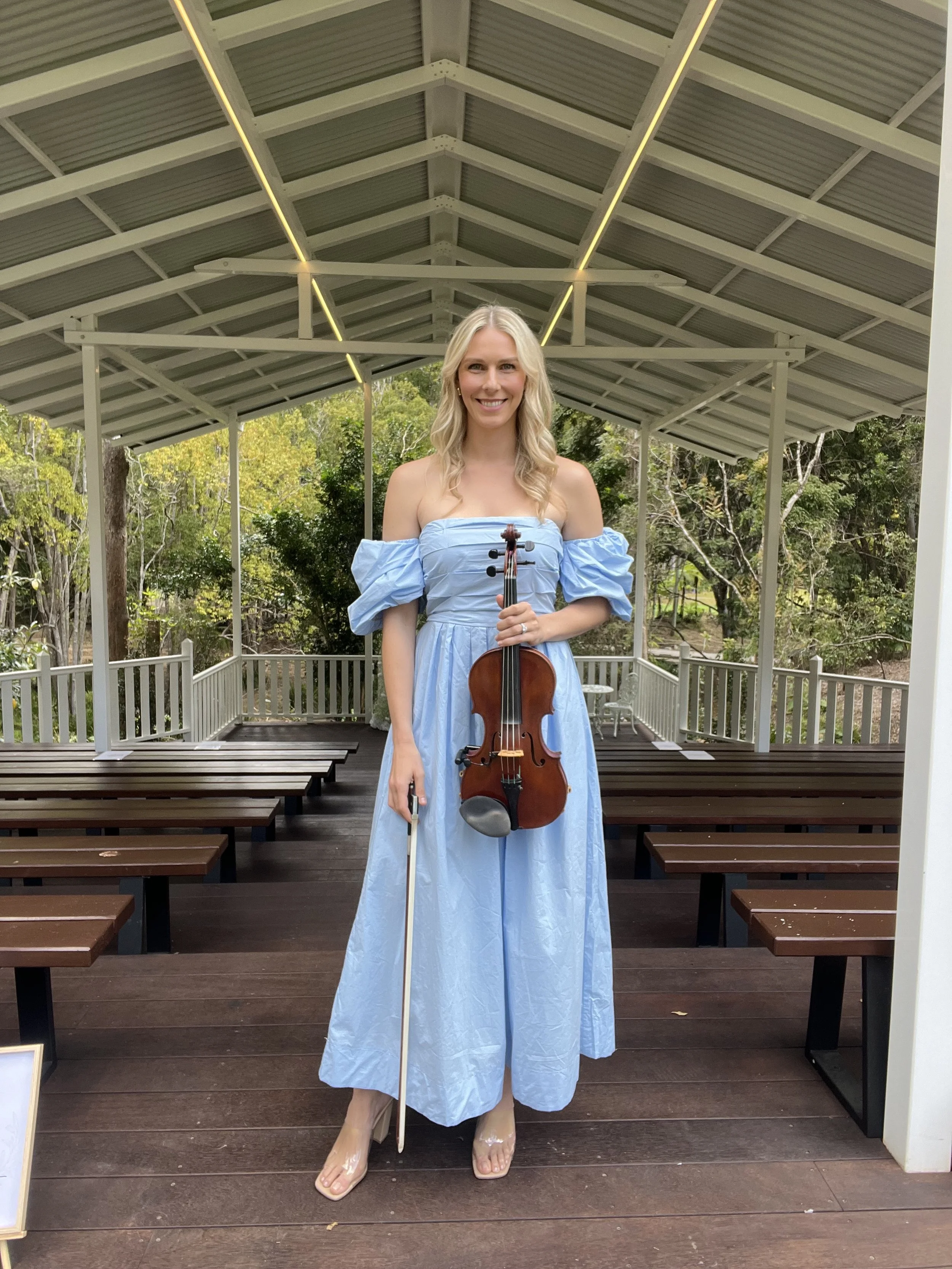 Lauren Bradnam Violinist in a light blue off-shoulder dress holding a violin and bow, standing on a wooden stage under a metal roof with trees in the background.
