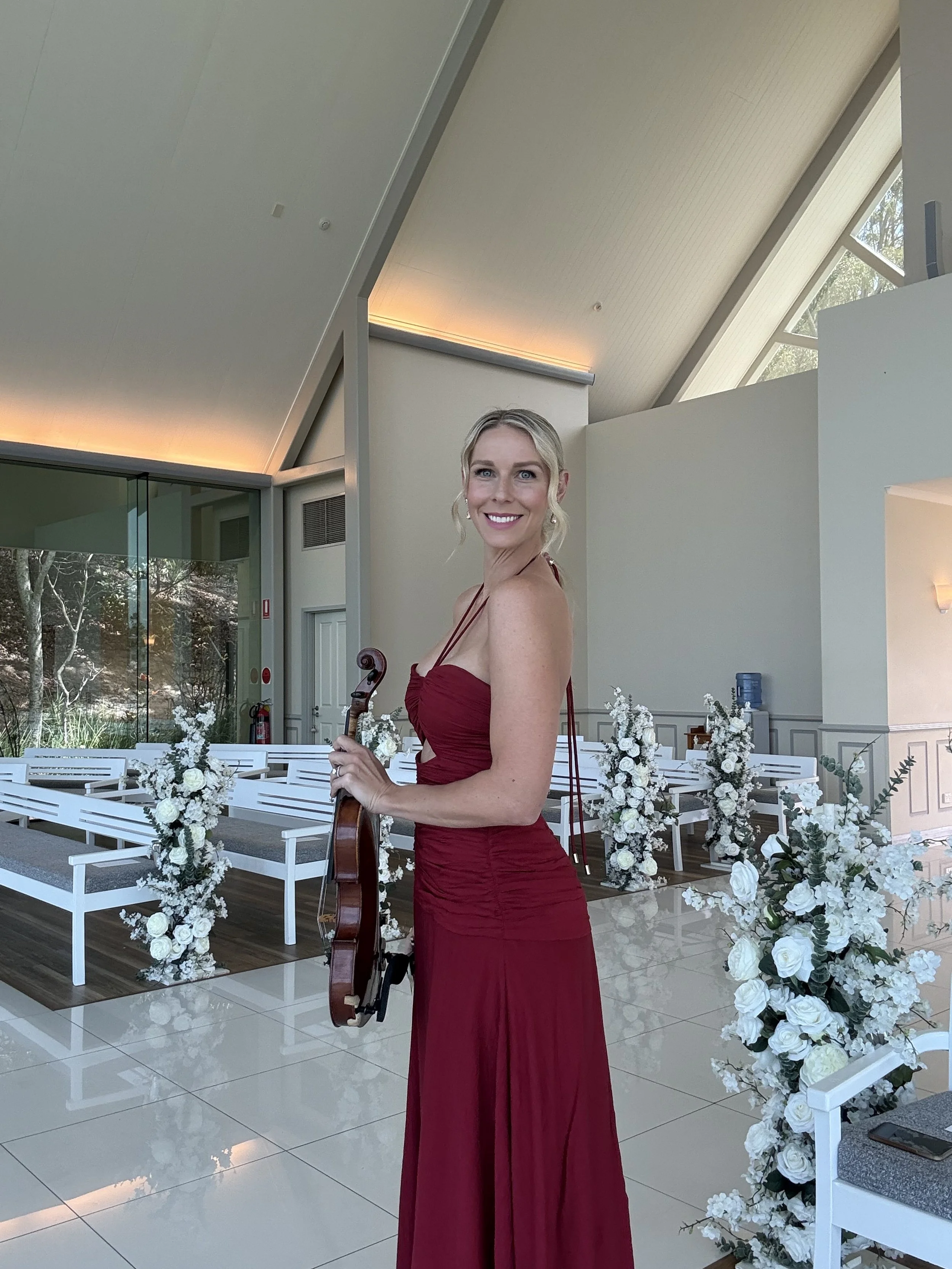 A woman in a strapless red dress holding a violin standing inside a modern decorated venue with white floral arrangements and benches.