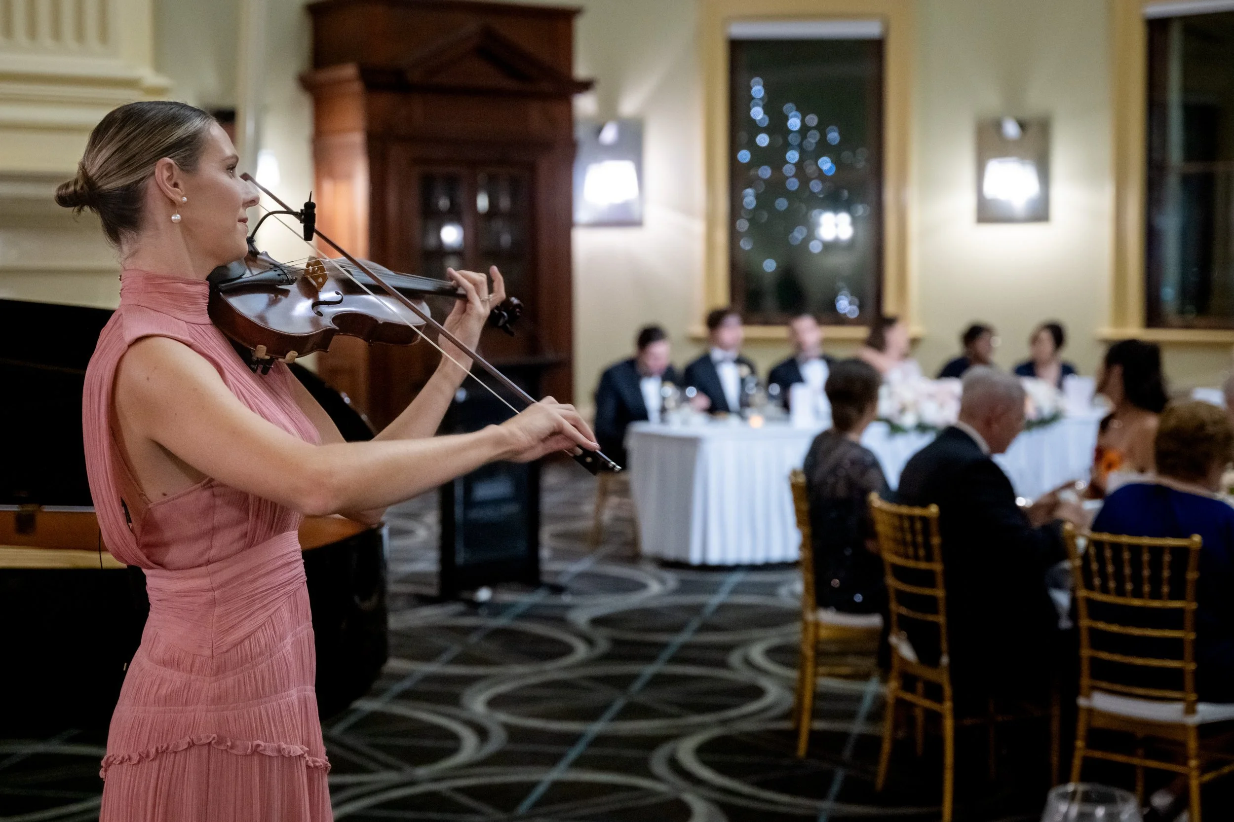 Lauren Bradnam Violinist in a pink dress playing the violin at a formal dinner event with people seated at tables in a decorated banquet hall. Playing at Customs House Brisbane.