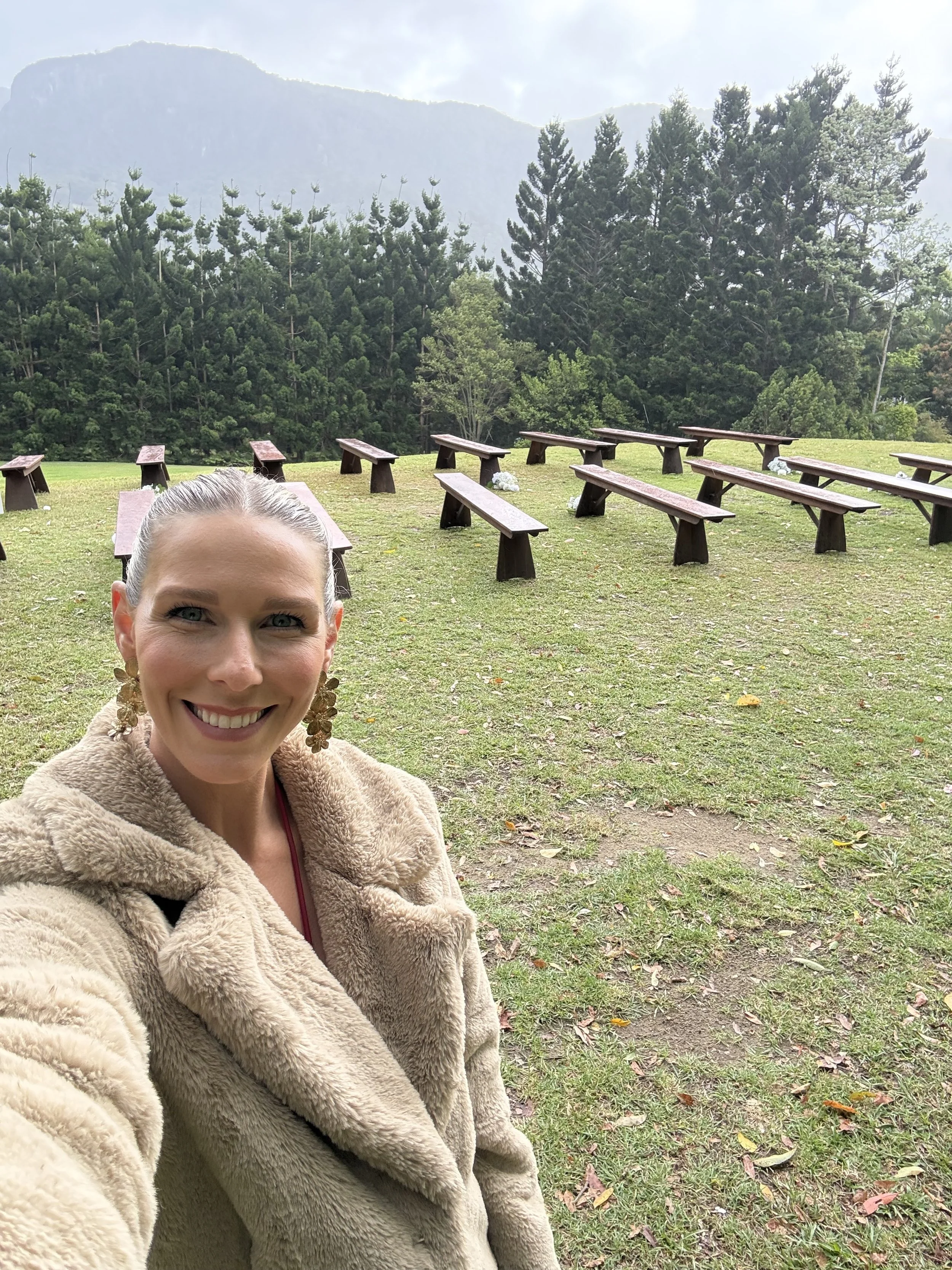 A woman taking a selfie outdoors in a grassy area with wooden benches arranged in rows behind her, surrounded by trees and mountains in the distance.