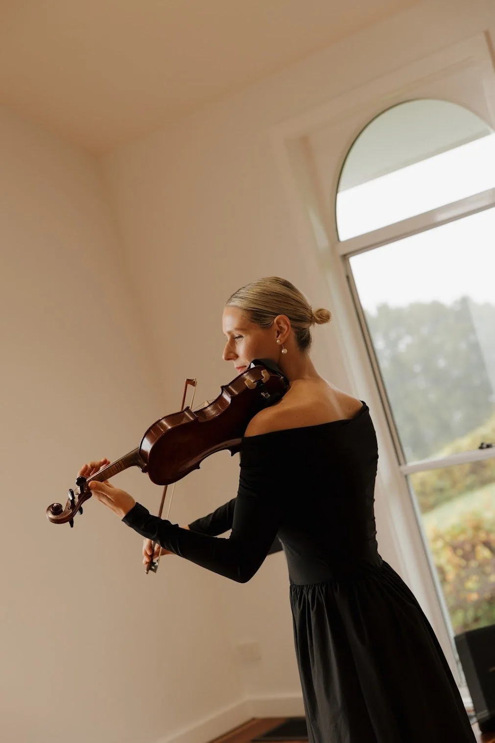 Lauren Bradnam Violinist playing the violin indoors near a large window, dressed in a black off-shoulder dress with pearl earrings at Summergrove Estate