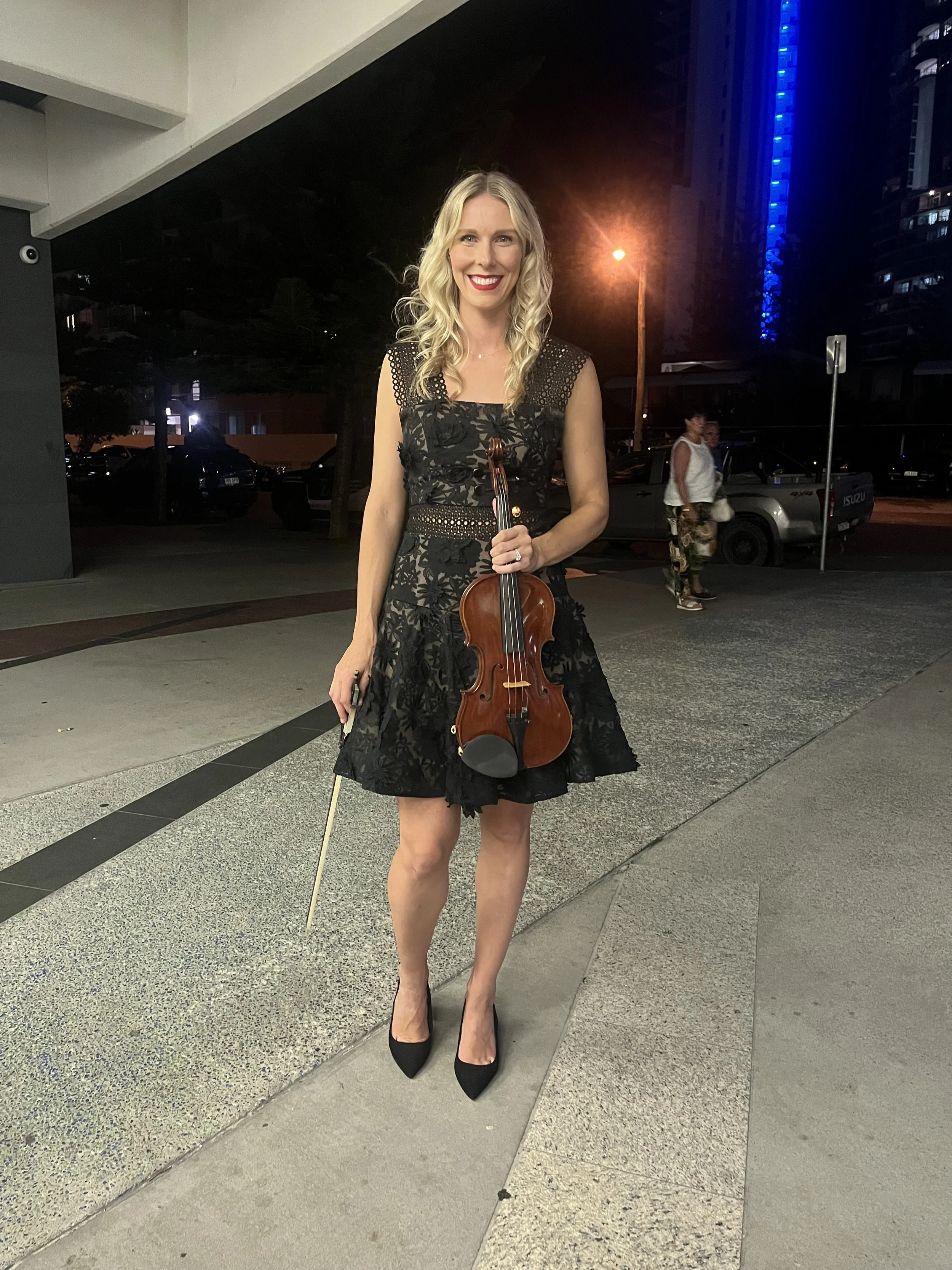 A woman in a black lace dress and high heels holding a violin and bow, standing outdoors at night on a city sidewalk with buildings and streetlights in the background.