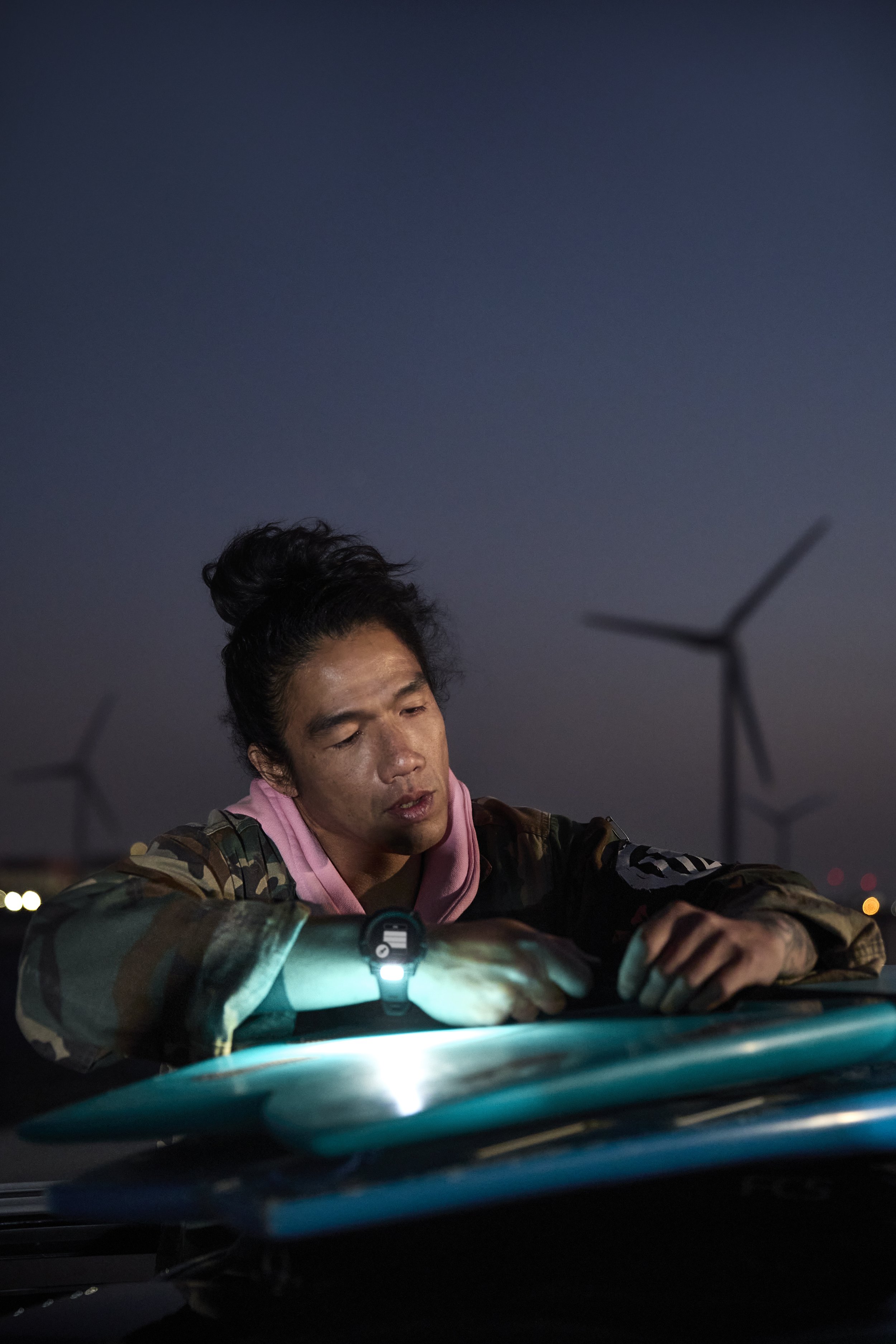 Person checking surfboards in low light near wind turbines.