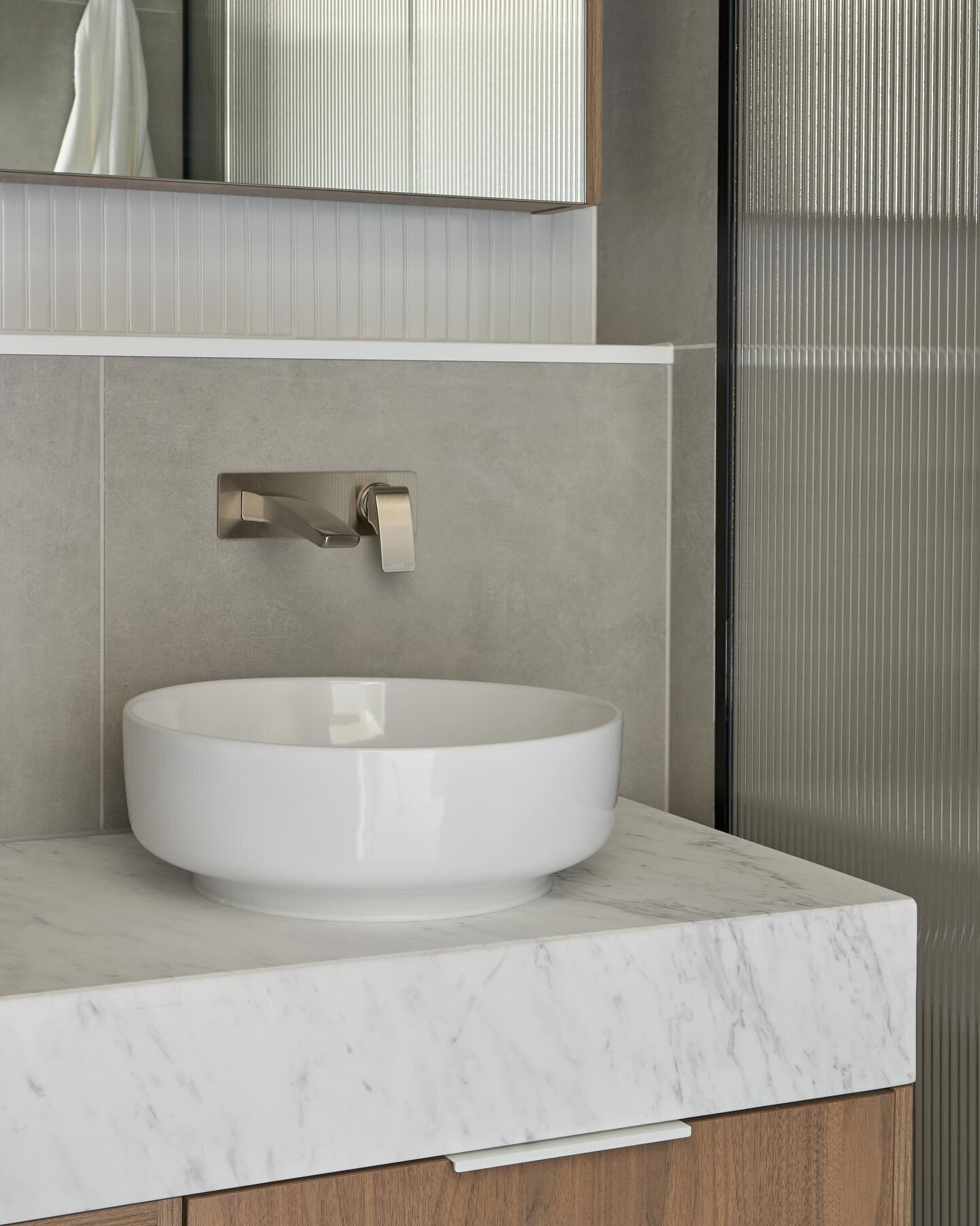 The use of Elba stone and textured timber laminate continued into the ensuite and bathroom for this custom apartment at Noir Northcote, the fluted glass continuing the theme set in the living room joinery. Lightly mottled grey tiles and Kit Kat tiles