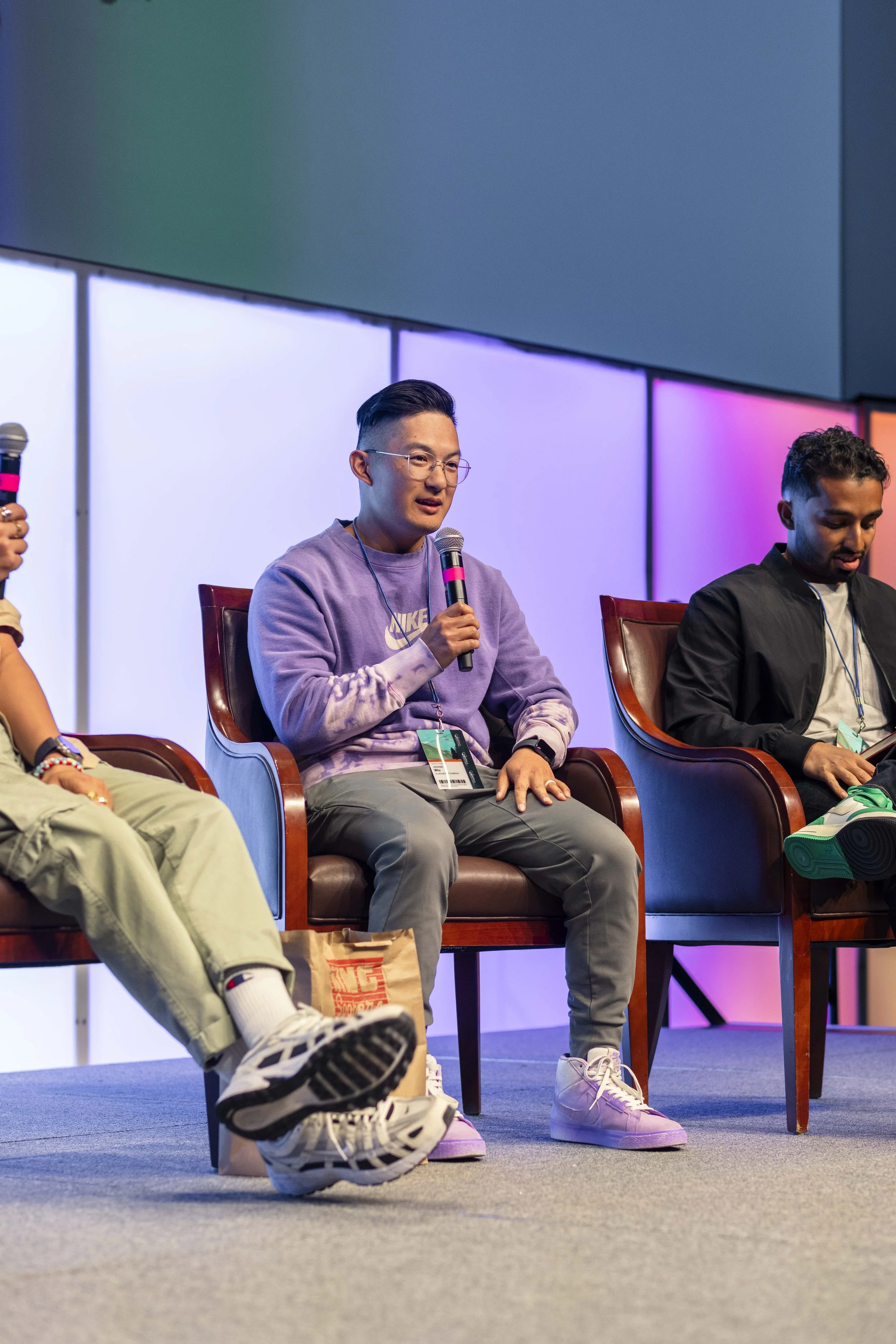 Three men sitting on stage in leather chairs during panel discussion at conference, one holding a microphone, with colorful background lighting.