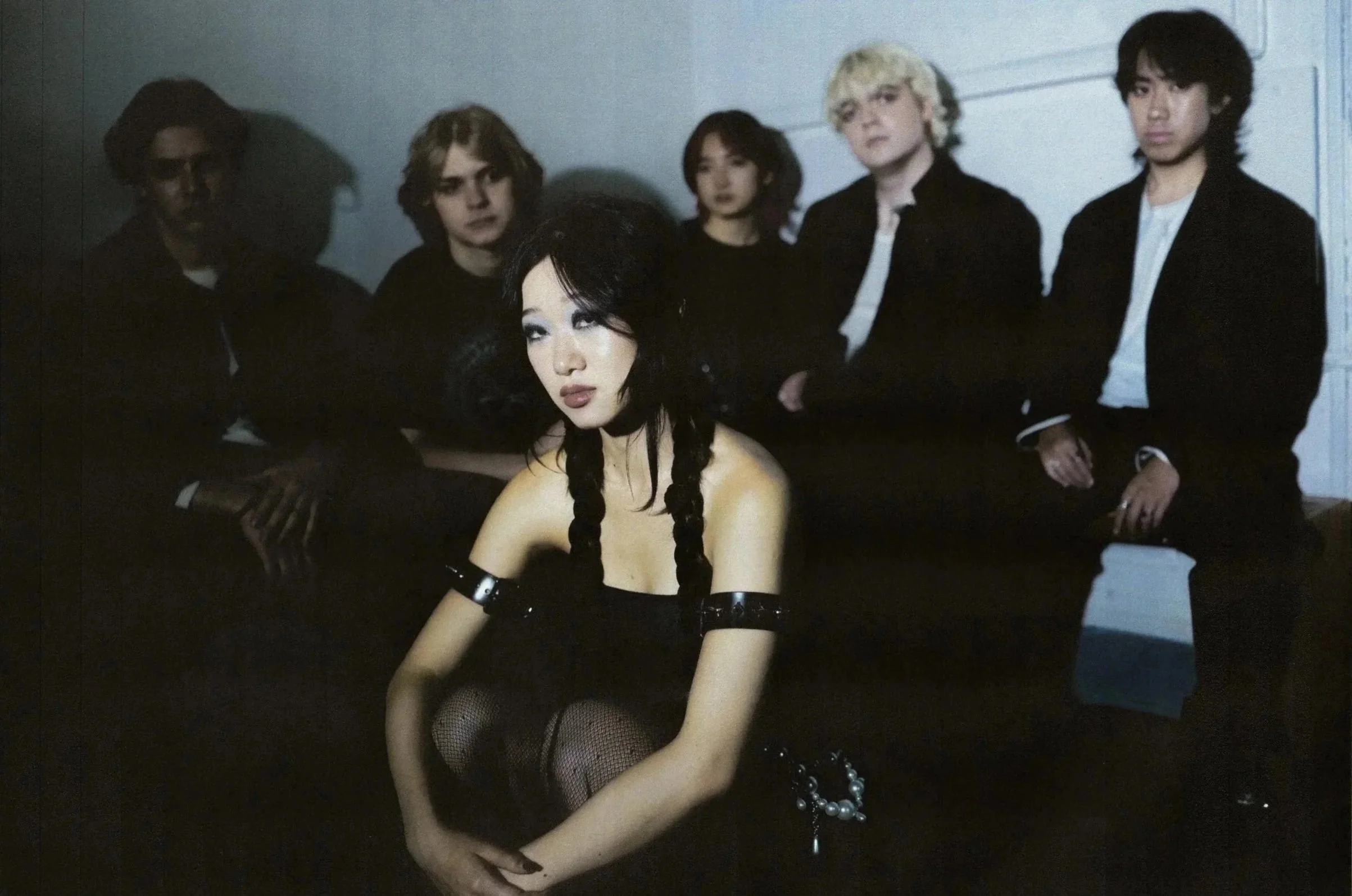 Group of five young adults, four men and one woman, in dark clothing, sitting against a plain wall, with the woman positioned in front, looking directly at the camera.