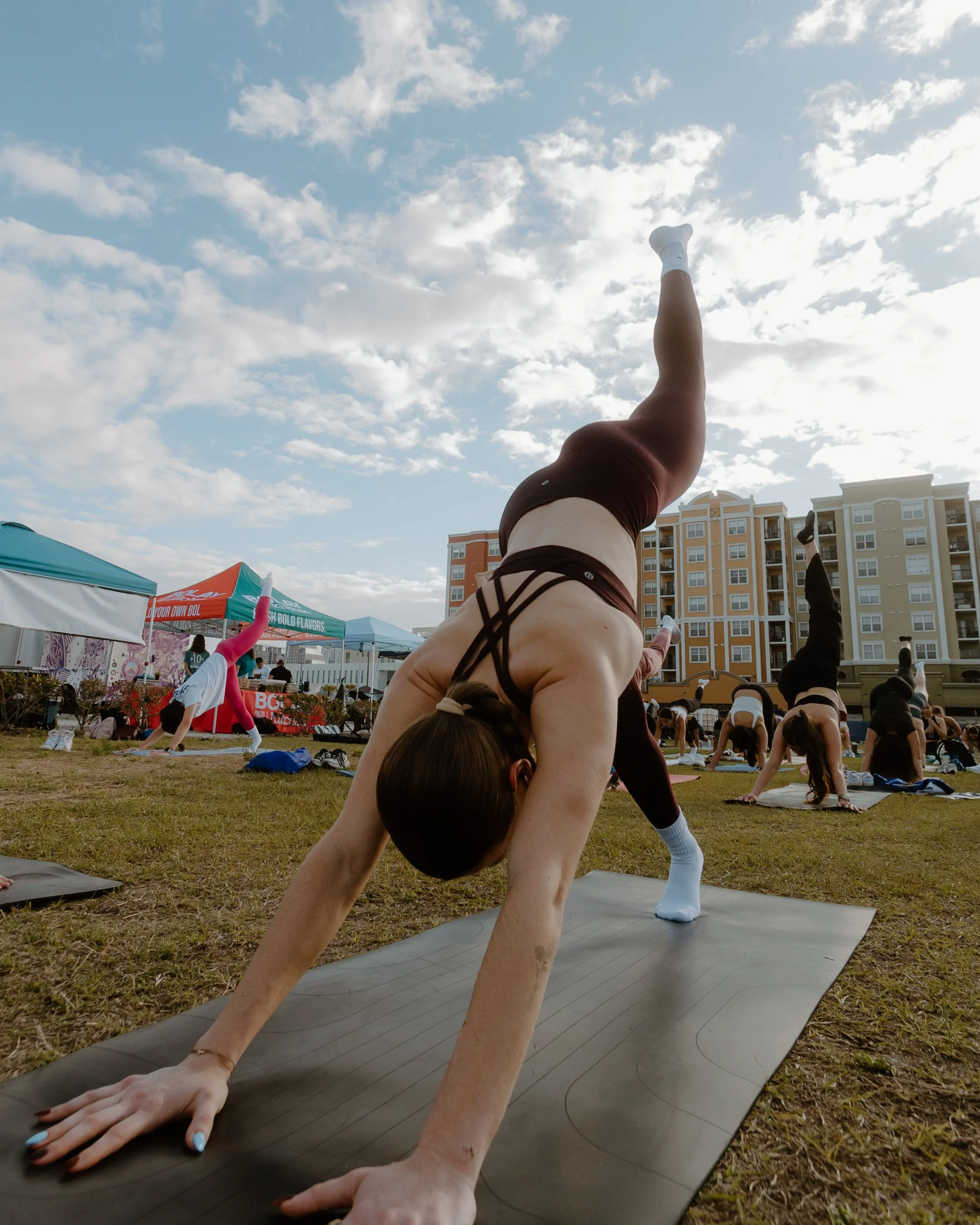 A woman practicing yoga outdoors on a black mat, stretching with her hands on the ground and one leg extended upward, surrounded by other participants in a yoga class on grass with tents and apartment buildings in the background.