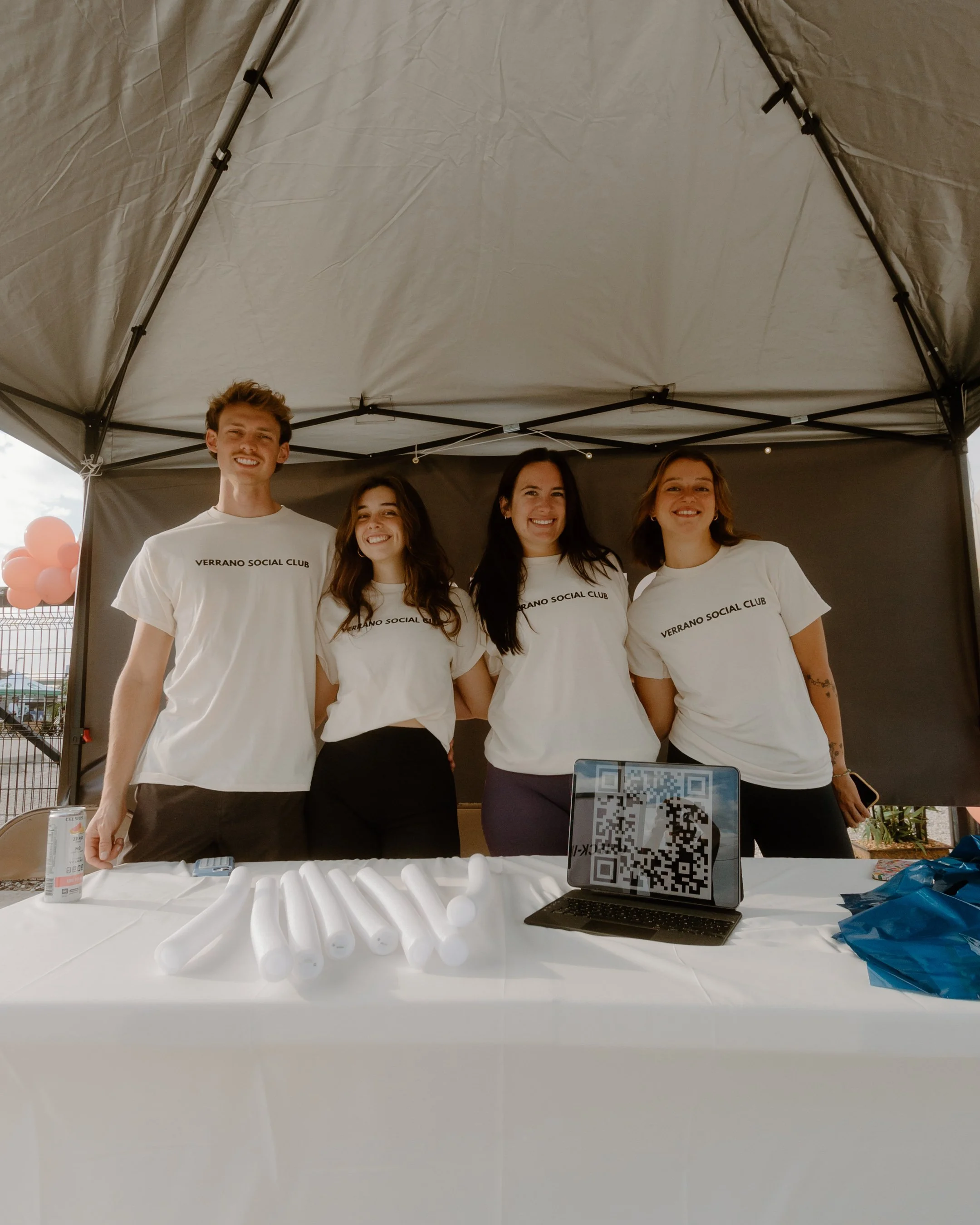 Four young people standing behind a table under a canopy, all wearing white t-shirts with 'VERRANO SOCIAL CLUB' printed on them, smiling at the camera.