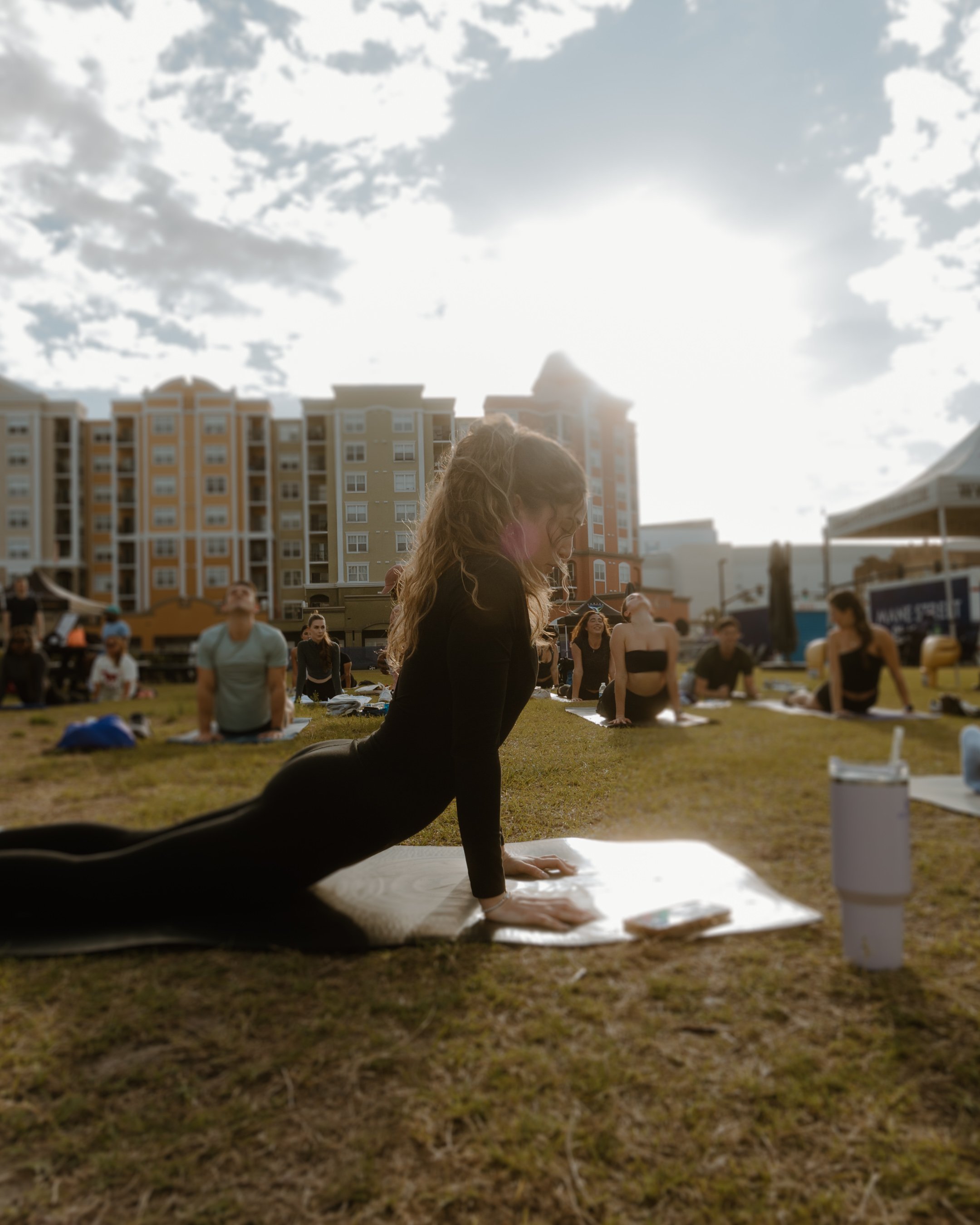 A group of people practicing yoga outdoors on a grassy area in front of tall apartment buildings. The focus is on a woman in black yoga attire performing the cobra pose with the sun shining behind her.