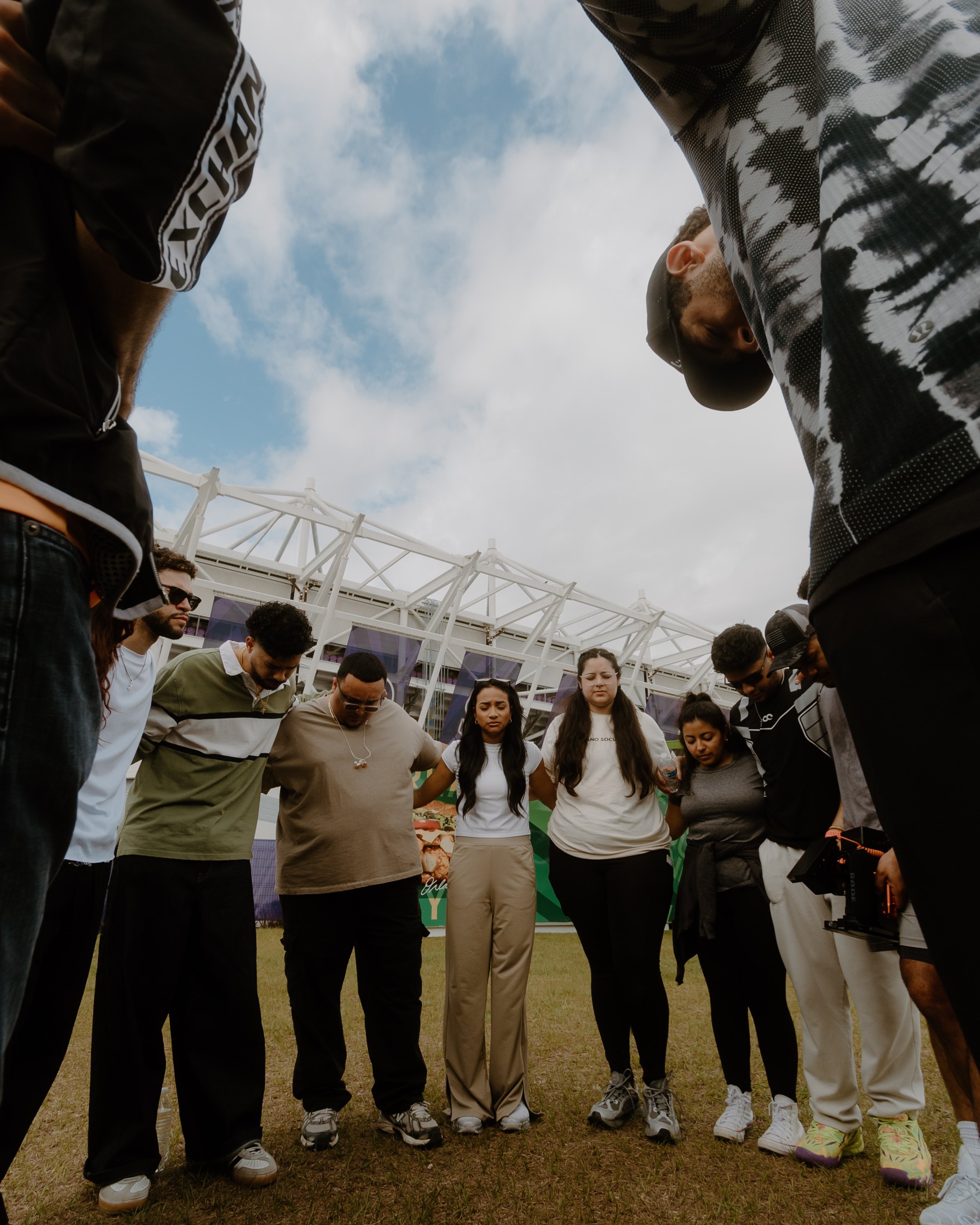 Group of people standing in a circle with heads bowed down, holding hands in an outdoor setting under cloudy sky