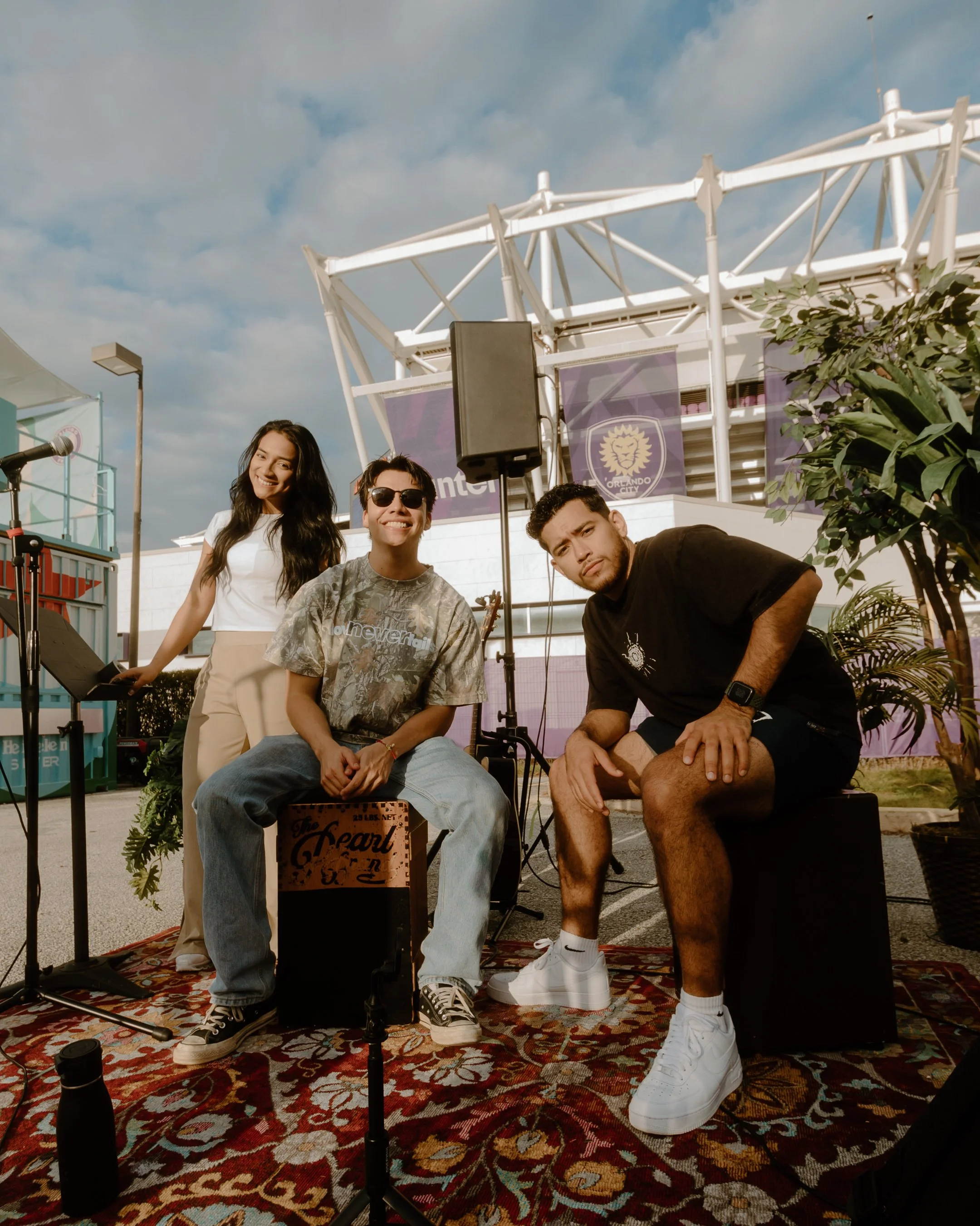 Three people sitting and standing in front of a microphone setup outdoors, with a stage structure and banners in the background, on a sunny day.