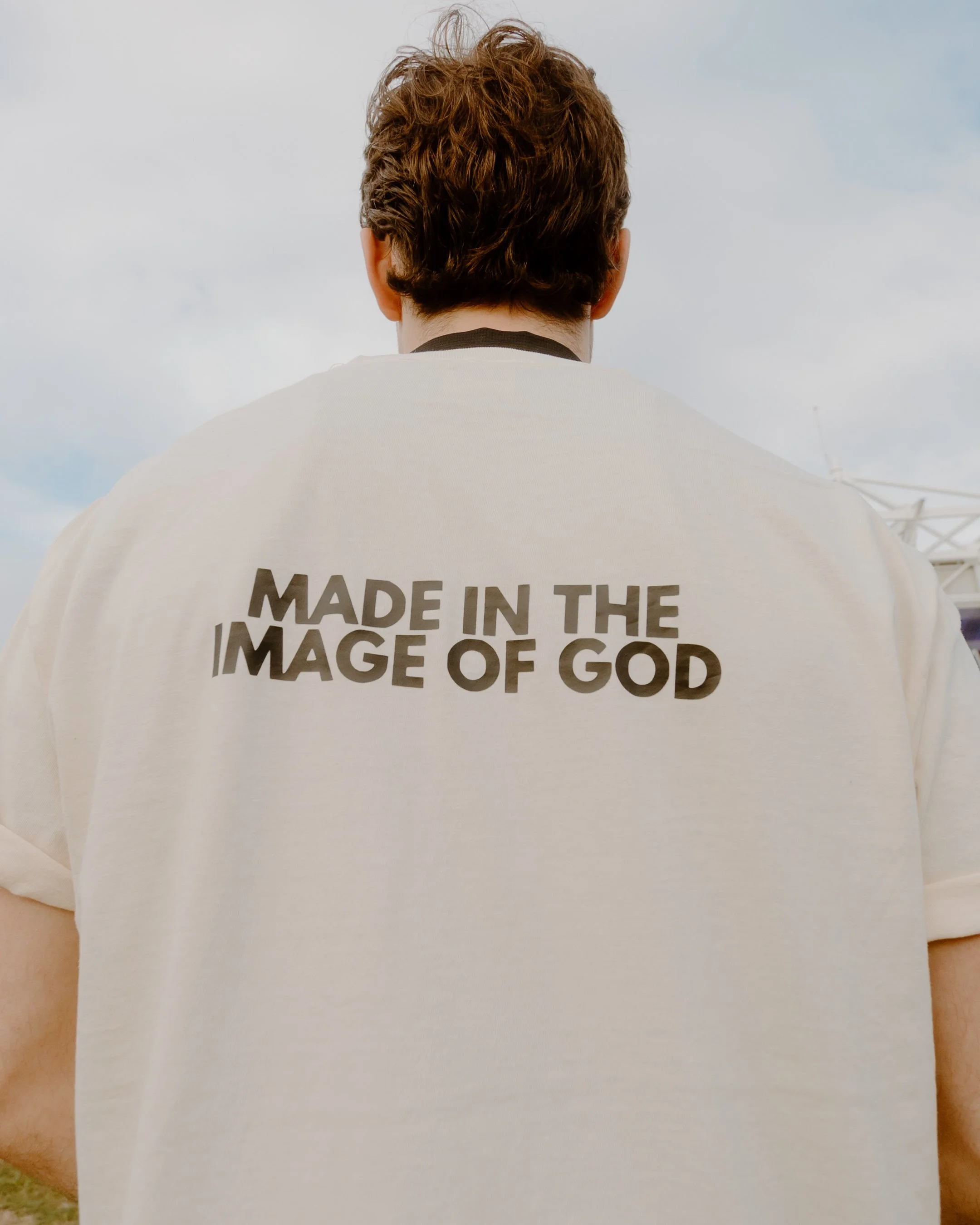 Back of a person wearing a white shirt with the words 'Made in the image of God' printed on it, outdoors on a cloudy day.