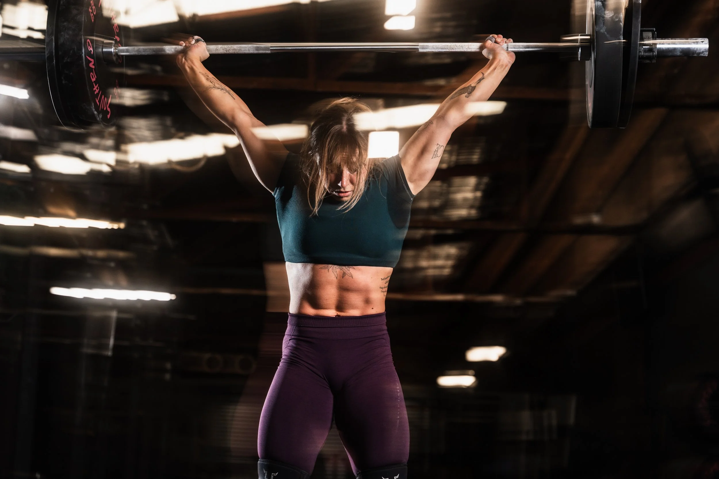 A woman with tattoos on her arms and torso lifting a barbell overhead in a gym, with motion blur indicating movement.