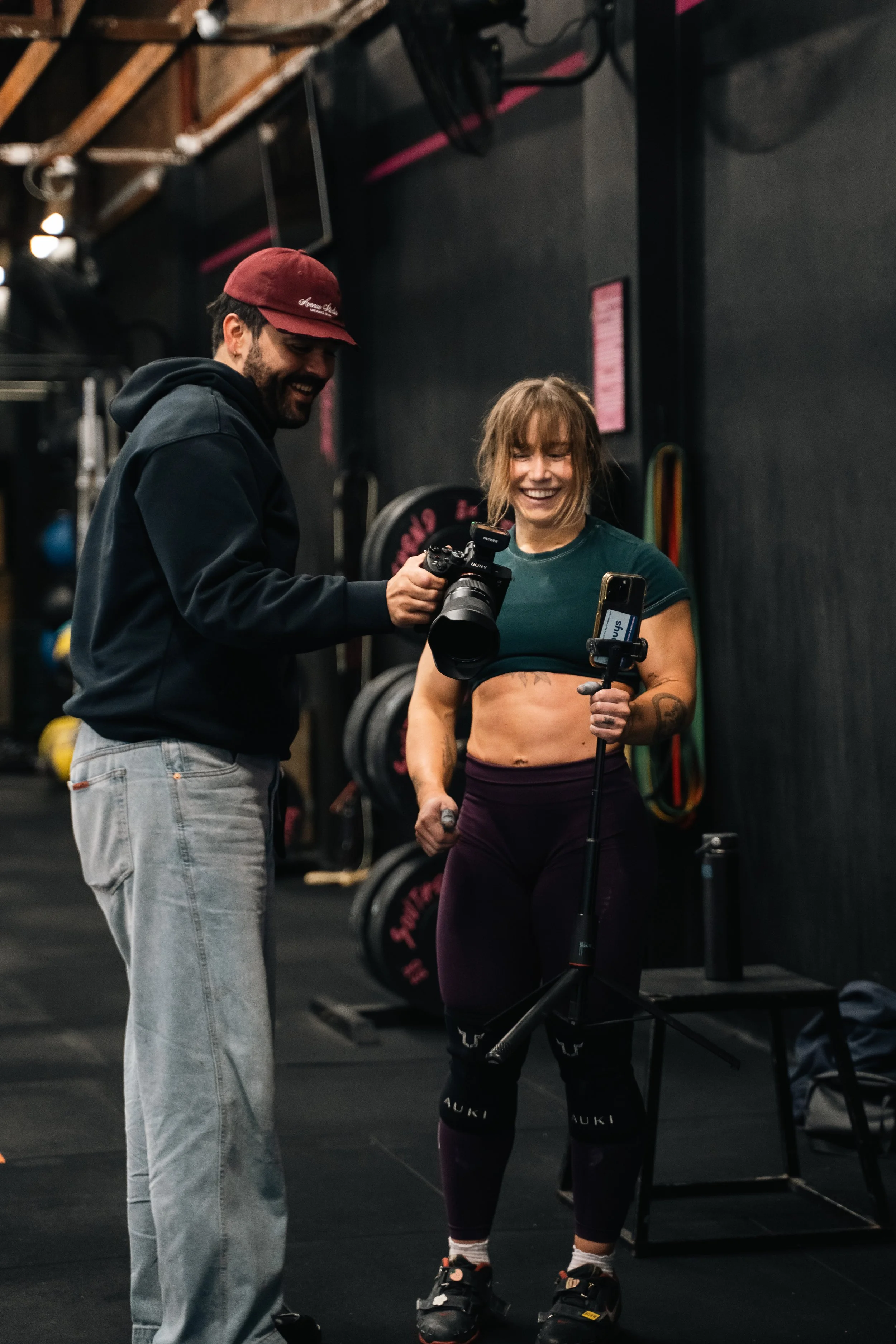 A woman in workout clothes holding a selfie stick and a dumbbell, smiling as a man with a camera stands next to her, in a gym setting with weights and equipment in the background.