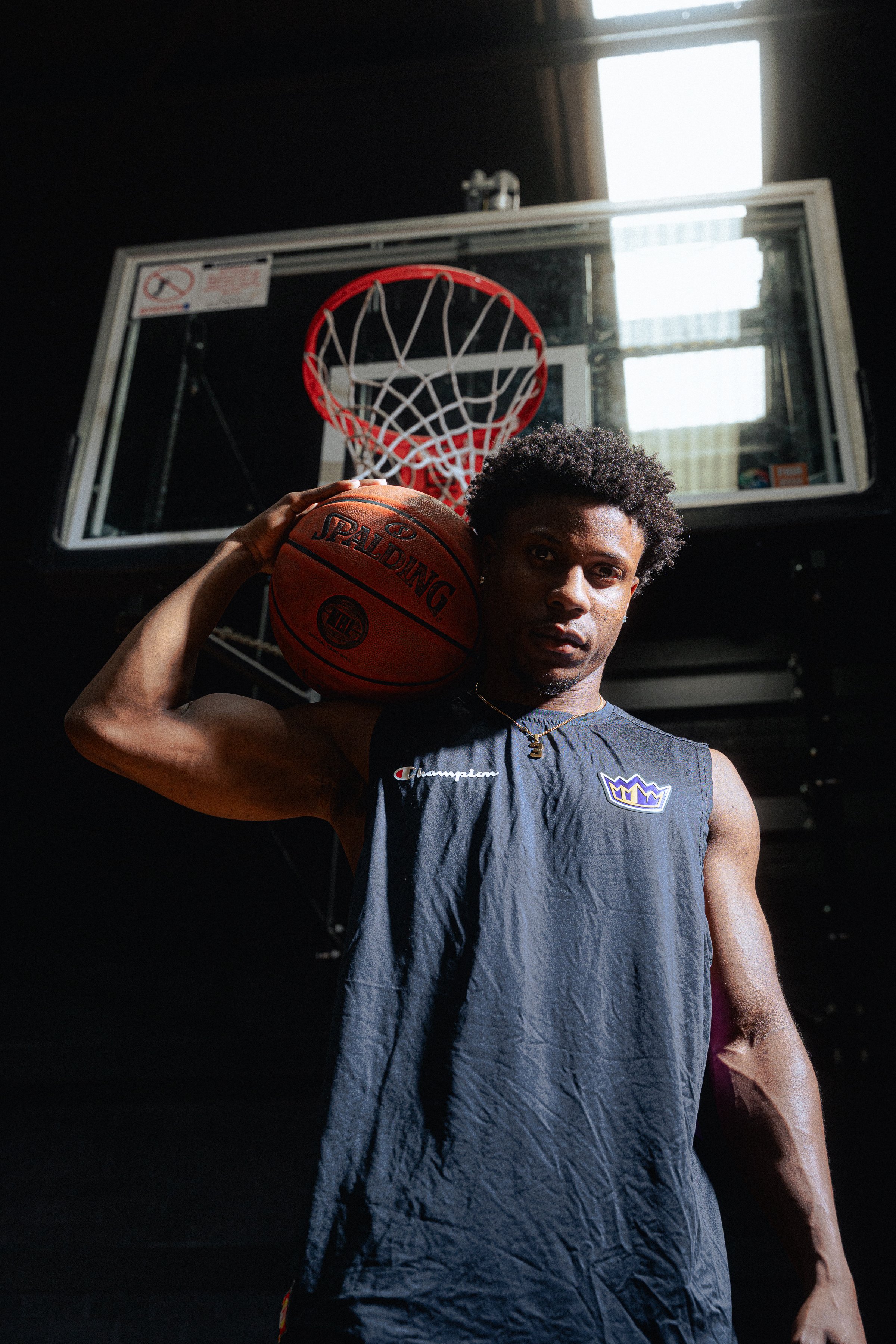 Basketball player holding a Spalding basketball near a hoop, wearing a Champion sleeveless shirt, in an indoor court.