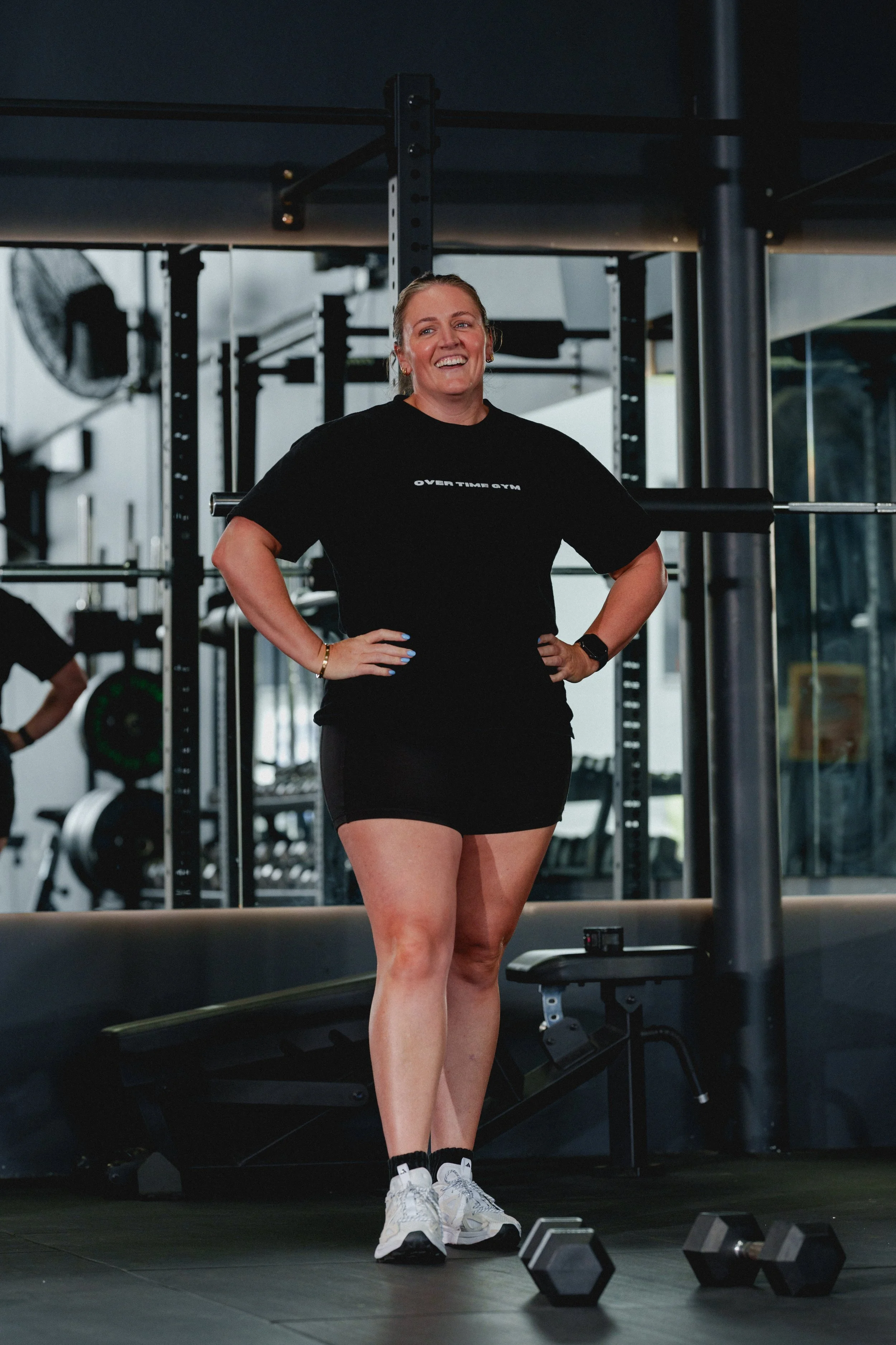 A woman standing in a gym, smiling, wearing a black t-shirt, black shorts, and white sneakers, with dumbbells on the floor nearby.