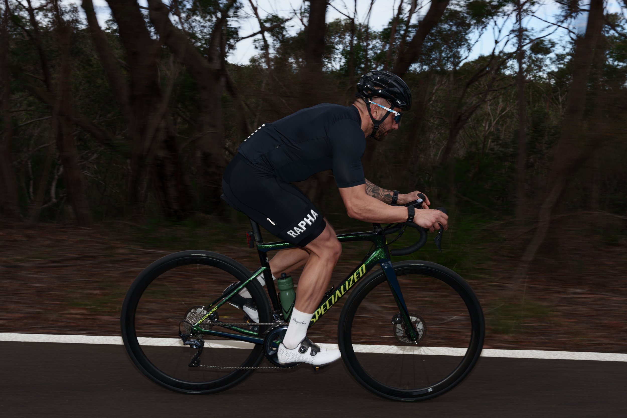 A man wearing a black helmet, sunglasses, a black cycling jersey, and black Rapha shorts riding a green Specialized road bike on a paved road with a forest background.