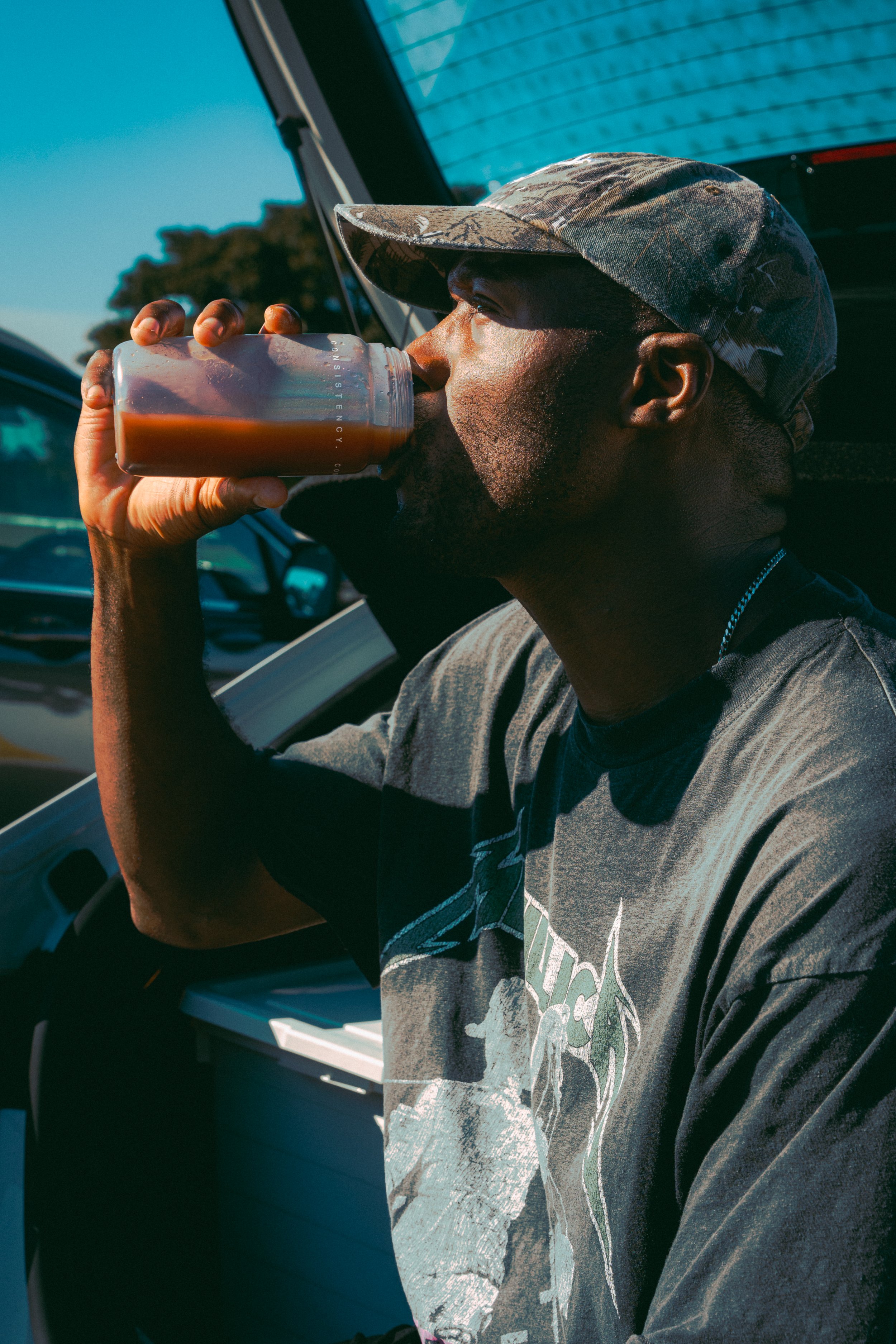 A man in a camo baseball cap and graphic t-shirt drinks an iced coffee from a clear plastic cup while sitting inside a vehicle.