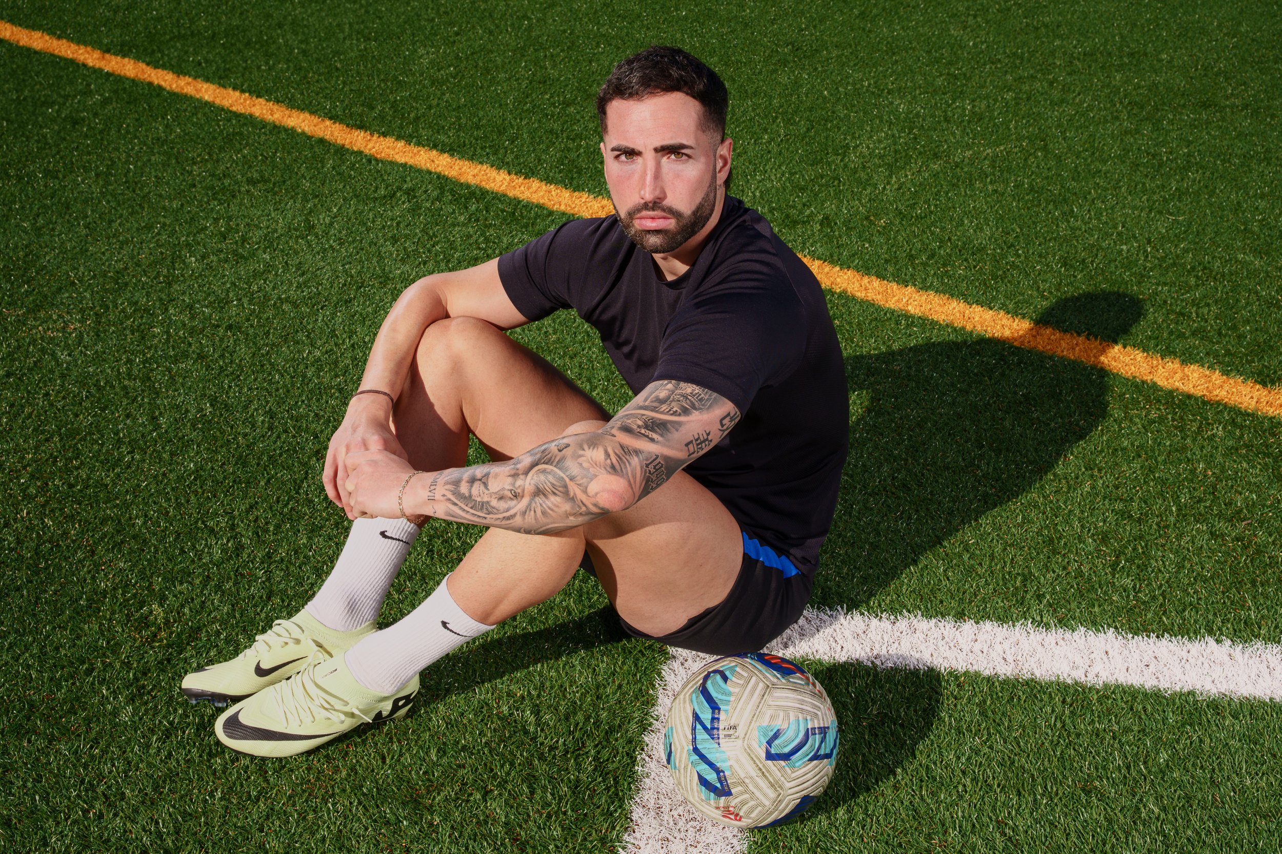 A man with short dark hair and tattoos on his right arm sitting on a green soccer field with a soccer ball beside him, wearing a black t-shirt, black shorts with blue accents, white socks, and yellow Nike soccer cleats.