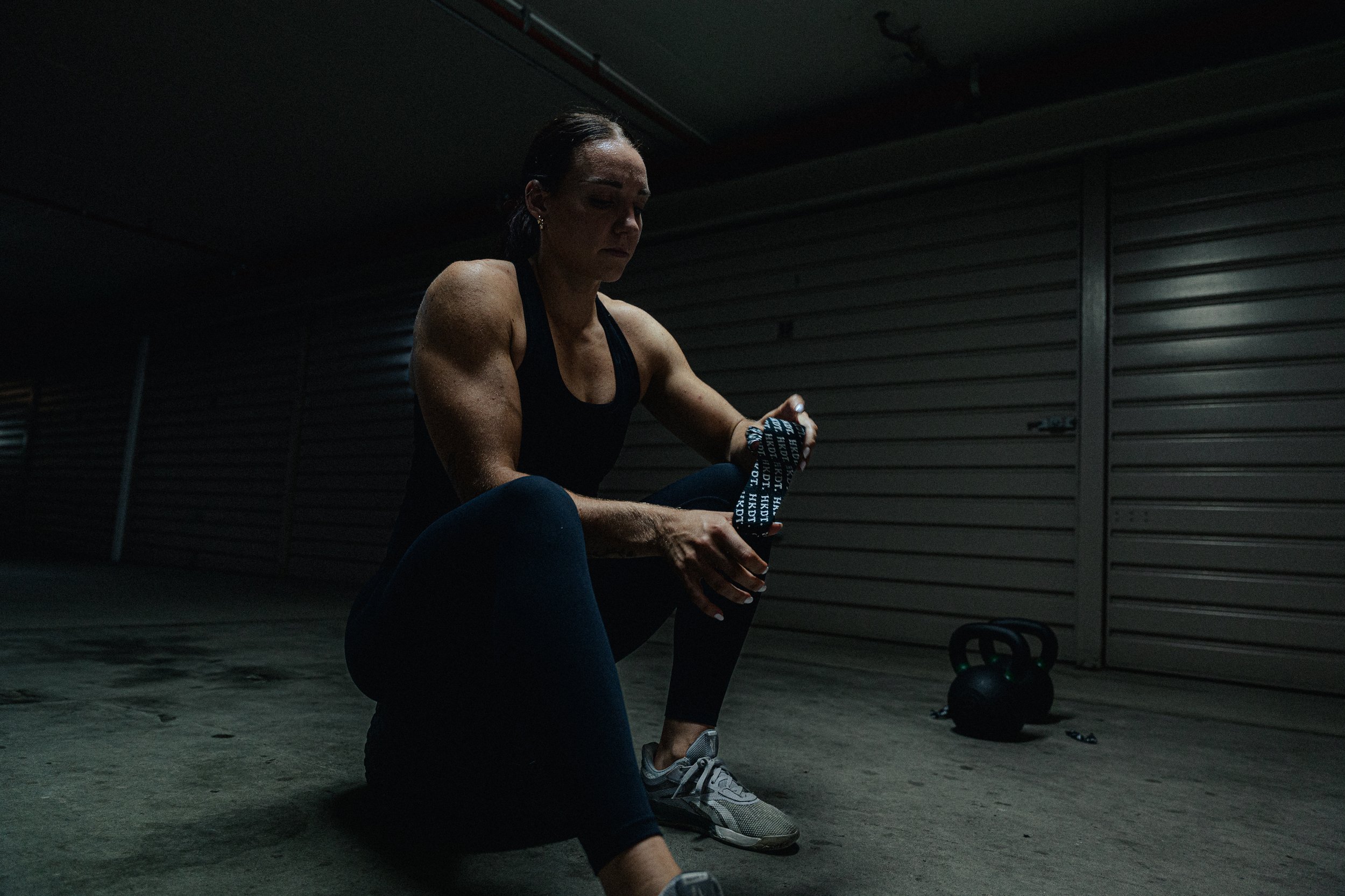 Person in gym attire sitting in a dimly lit garage with kettlebells nearby, holding a shaker bottle.