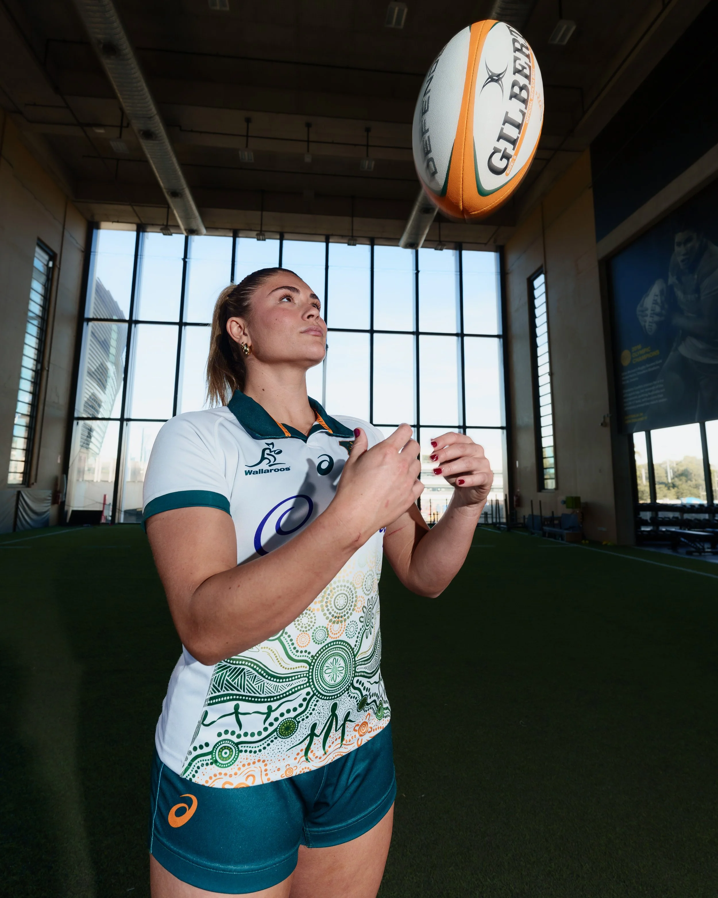A female athlete in a graphic sports uniform standing indoors, preparing to catch a Gilbert branded volleyball in a gym with large windows.