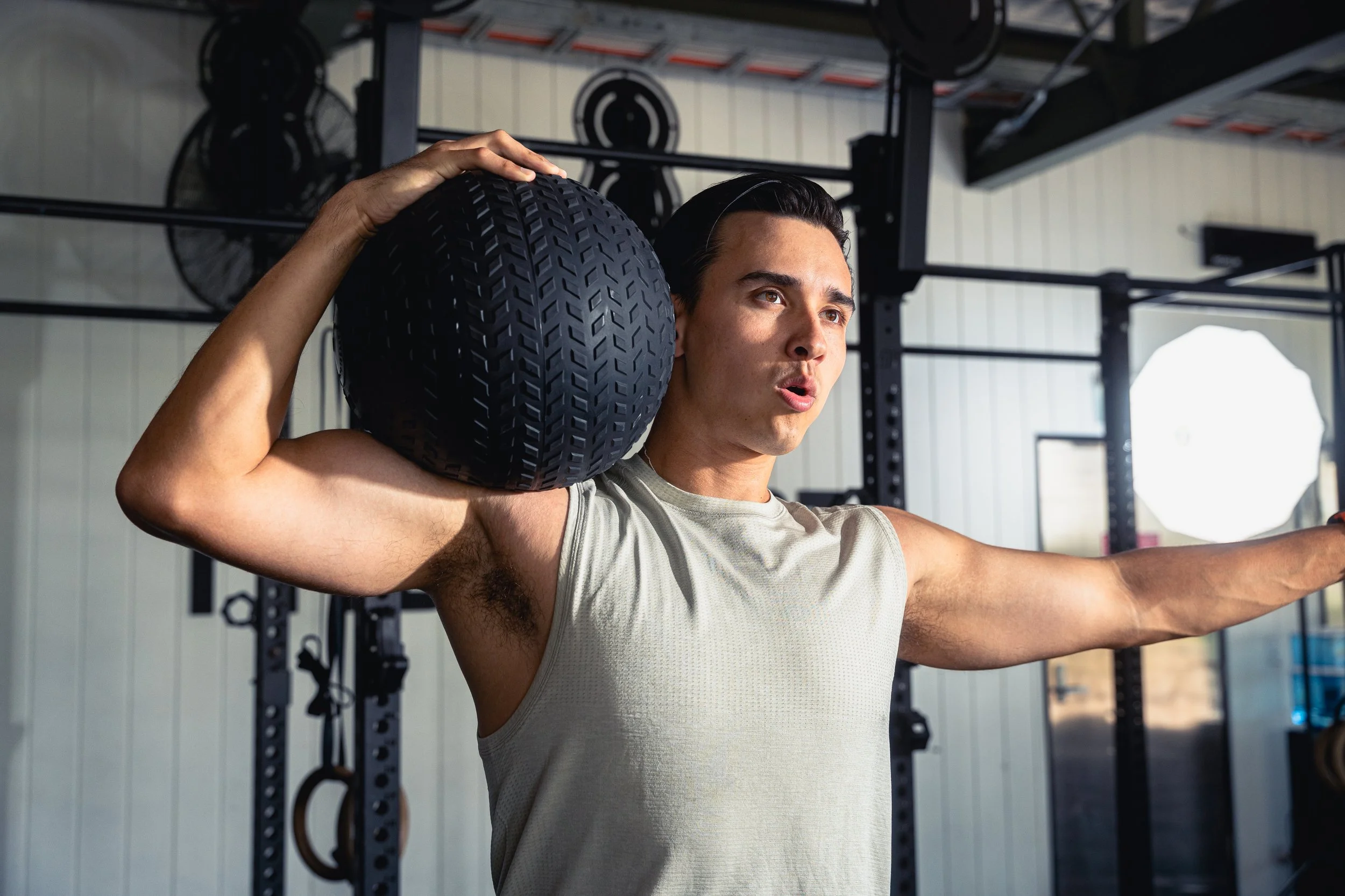 A man in a gym hoisting a black medicine ball on his shoulder, with his other arm extended outward, surrounded by gym equipment and a white wall.