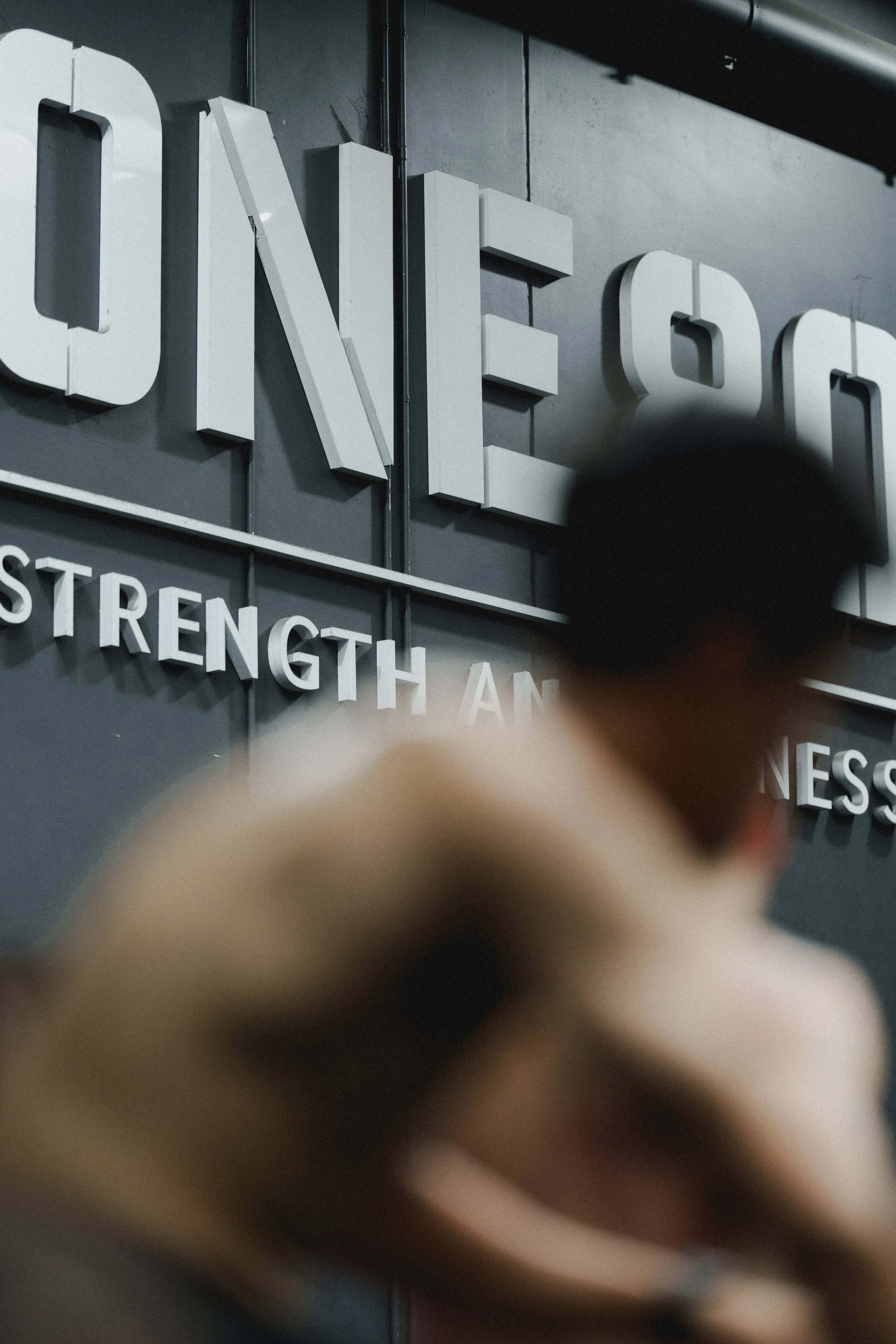 Close-up of a wall with large 3D letters spelling 'ONE' and partially visible words 'STRENGTH AND' in a gym or fitness center, with a blurred person in the foreground.
