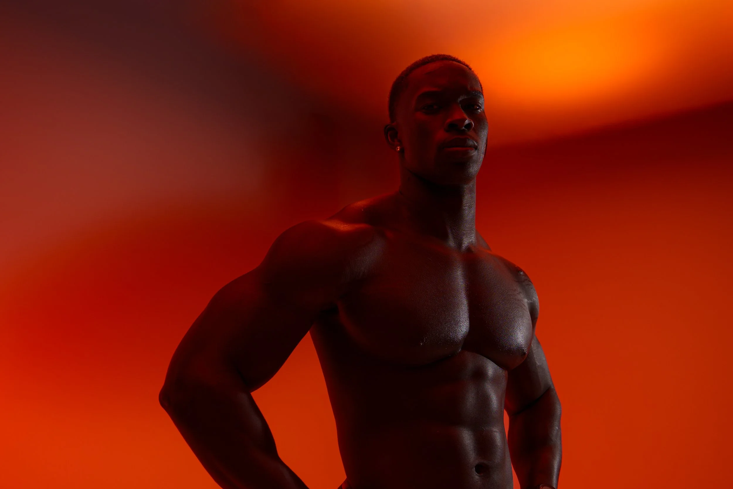 A shirtless man with muscular build standing against a vibrant orange background, looking confidently at the camera.