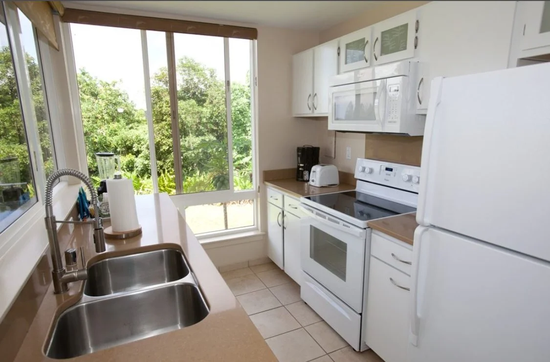 A small kitchen with white cabinets, a white refrigerator, white oven, microwave, toaster, coffee maker, and a window overlooking a green outdoor area.