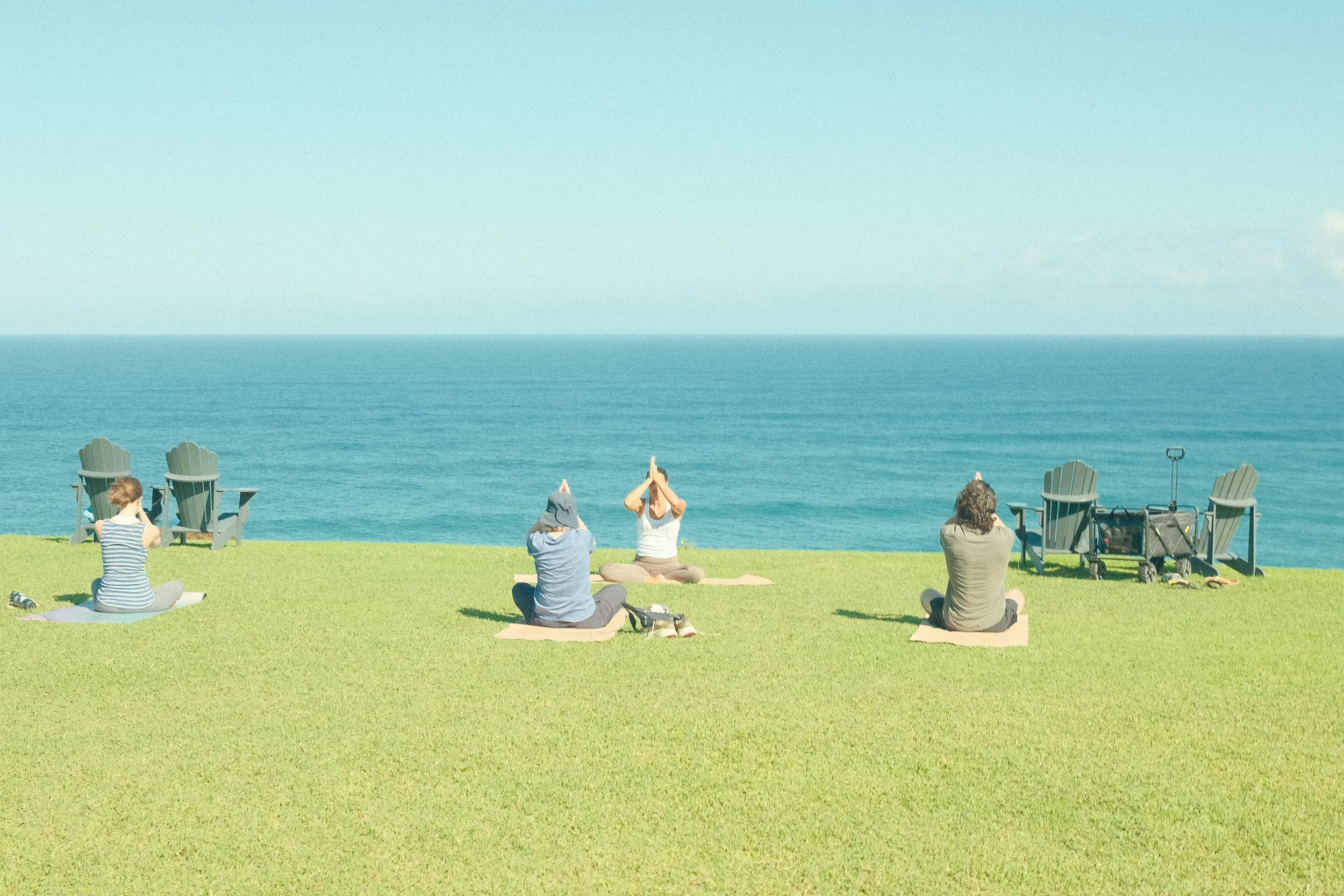 Four people practicing yoga on a grassy lawn overlooking the ocean, with two Adirondack chairs and a stroller on the right side.