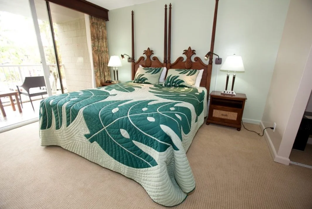 A bedroom with a four-poster bed with wooden headboard, a patterned green and white quilt, two pillows, two bedside lamps, a small nightstand, a sliding glass door with curtain, and balcony furniture outside.