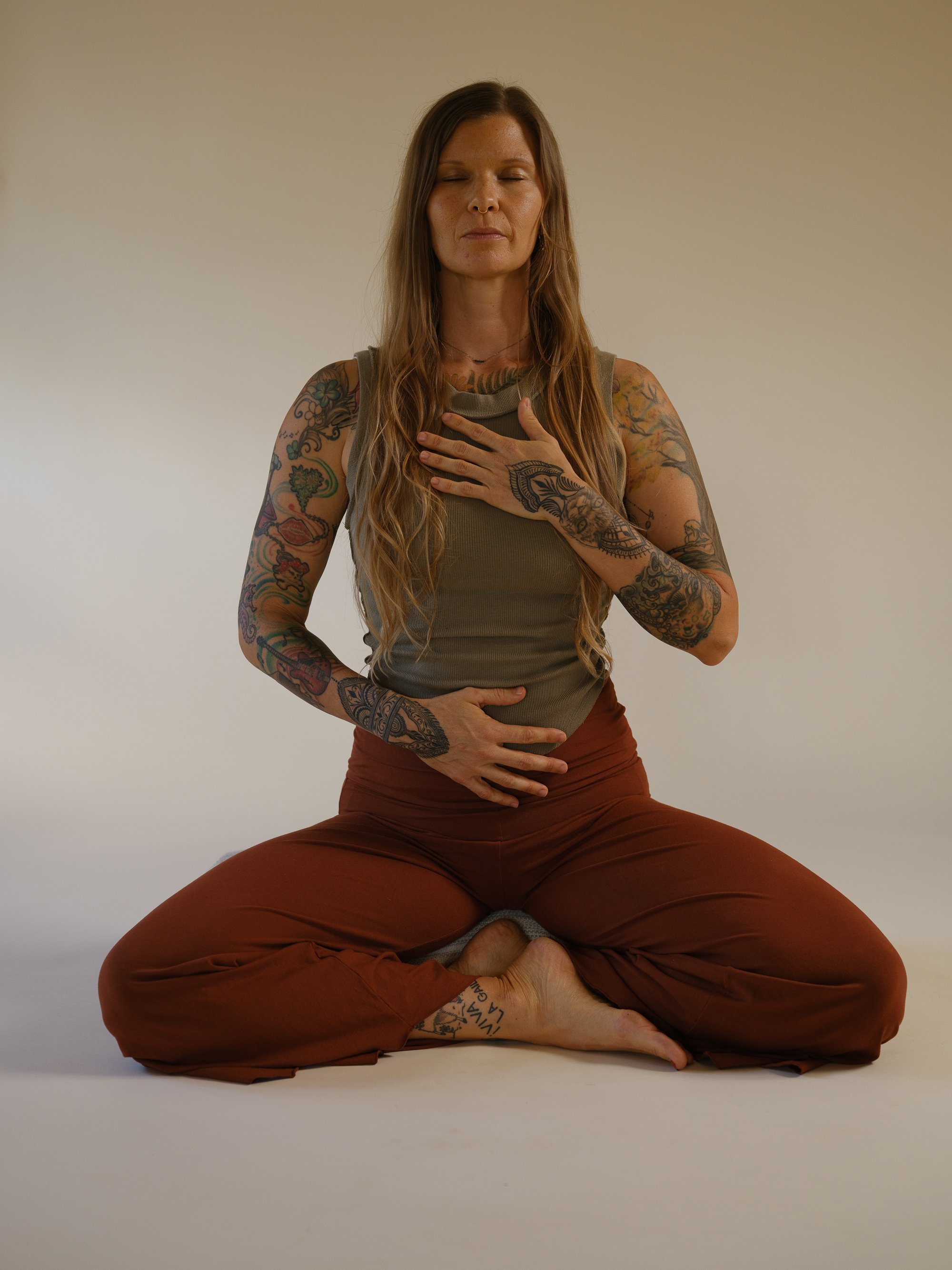 A woman wearing a brown sports bra and matching shorts practicing yoga in a wide lunge pose with her left hand on her left thigh and her right arm extended overhead, against a plain beige background.