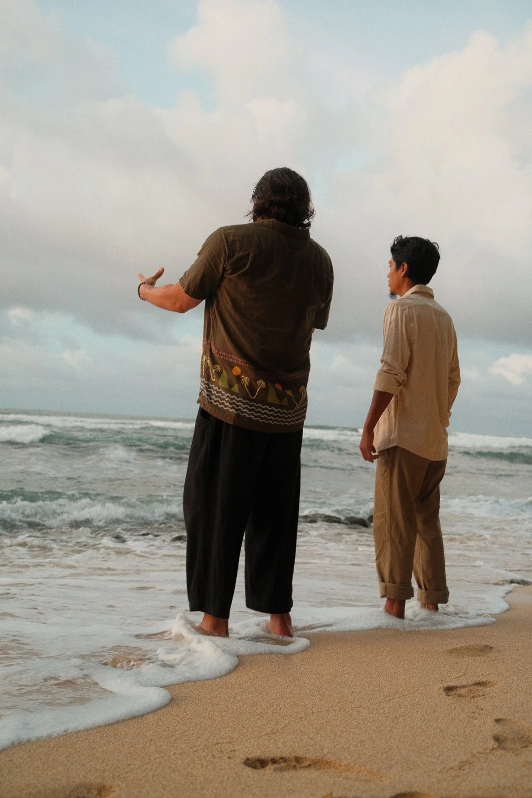 Two men standing barefoot on the beach near the ocean waves, one with long hair and wearing a dark shirt and pants, and the other with short, curly hair wearing a light-colored shirt and pants, looking out towards the water under a cloudy sky.