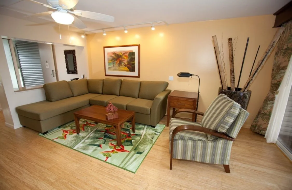 Living room with a beige sectional sofa, a wooden coffee table with pink coral decorations, a striped armchair, a small wooden side table with a black lamp, a colorful tropical rug, and a wall painting of a palm tree landscape.