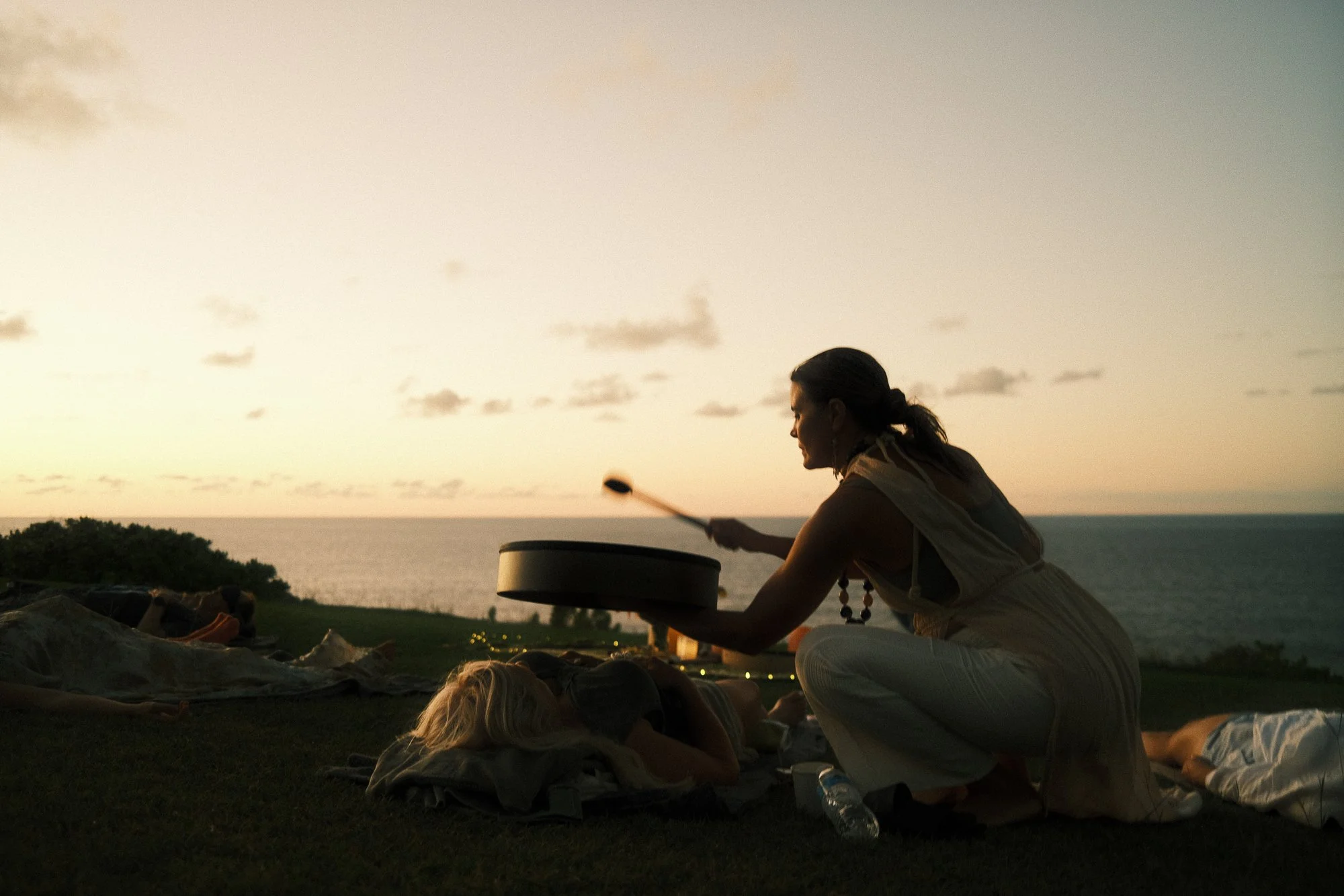 A woman crouched on the grass playing a hand drum during sunset by the ocean, with a person lying on the ground nearby.