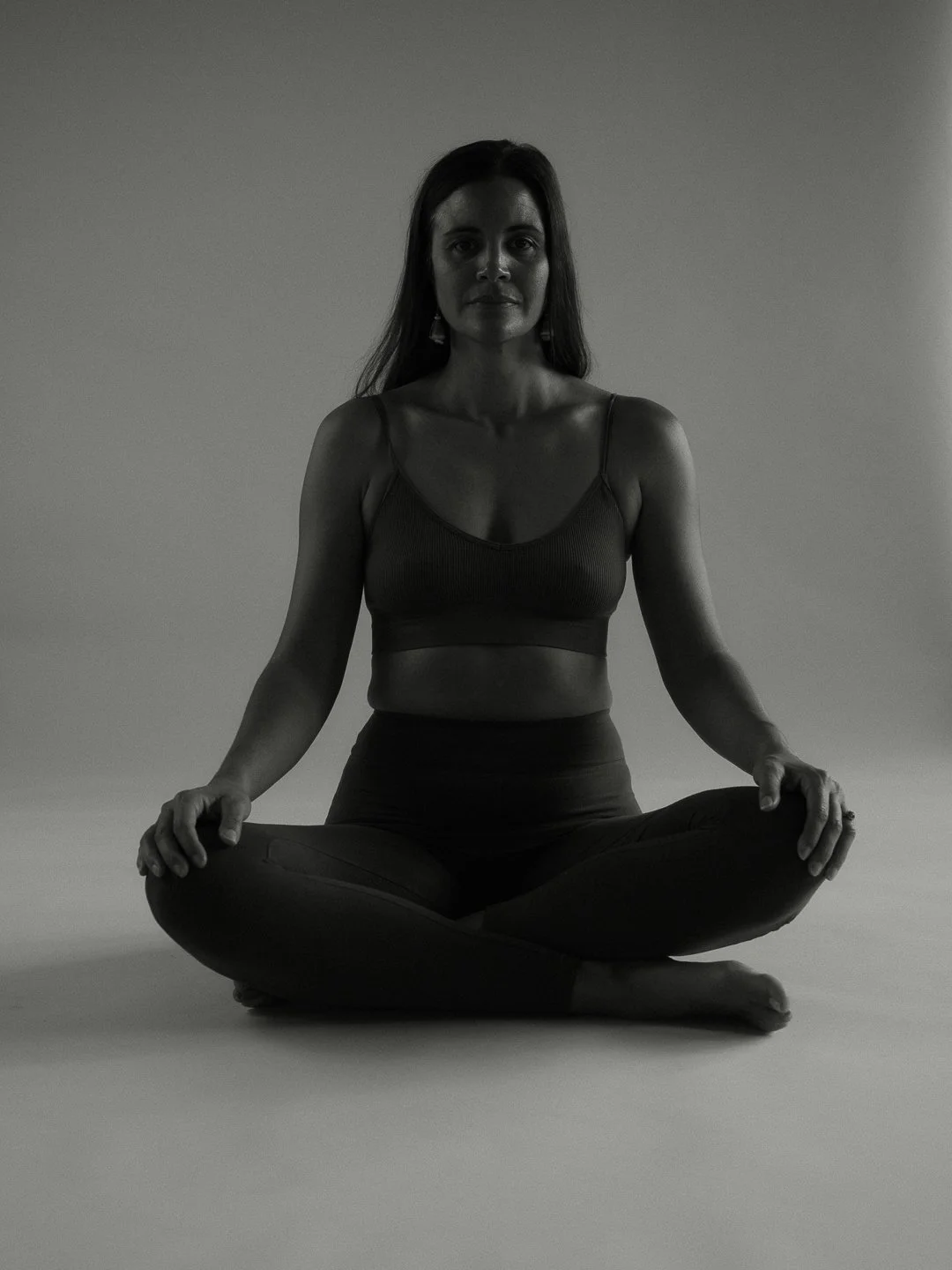 A woman sitting in a cross-legged yoga pose on the floor in front of a plain background.