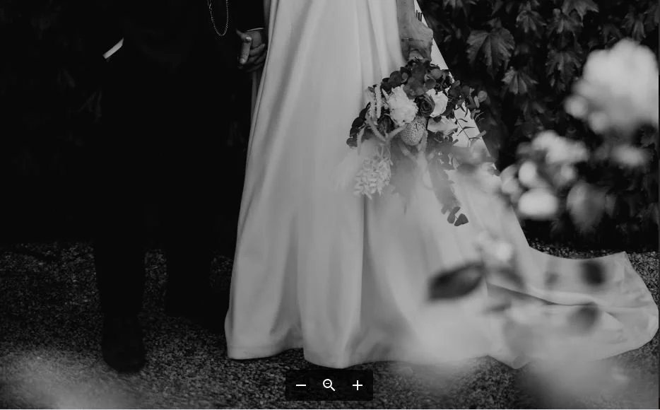 Black and white photo of a bride in a wedding dress holding a bouquet of flowers, standing beside a partially visible person in dark clothing, with leaves and flowers in the background.